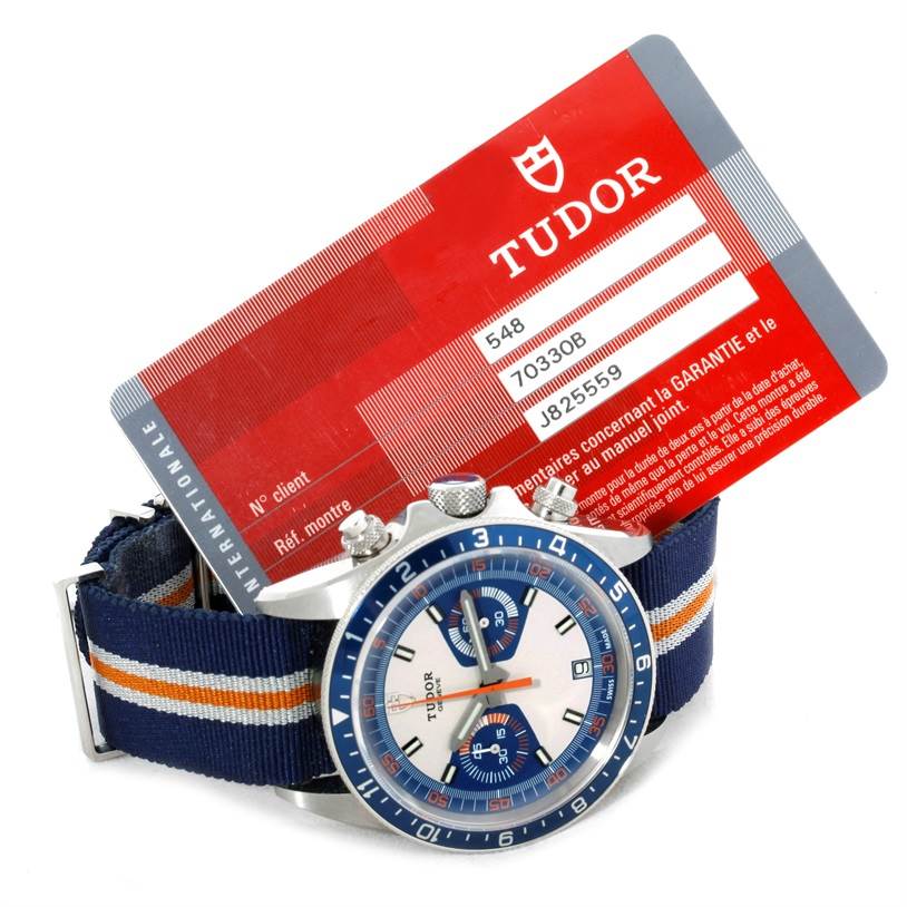 The image shows a frontal view of a Tudor Heritage Chrono 79220R Men's Stainless Steel Blue Dial 79220R Men's Stainless Steel Blue Dial watch with a fabric strap, accompanied by a Tudor warranty card in the background.