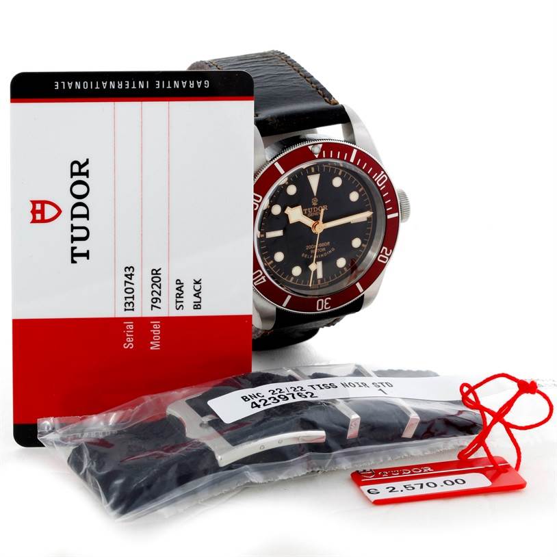 The Tudor Heritage Black Bay 79220R Men's Stainless Steel Black Dial 79220R Men's Stainless Steel Black Dial watch is shown from a front angle with accessories including a guarantee card and spare strap.