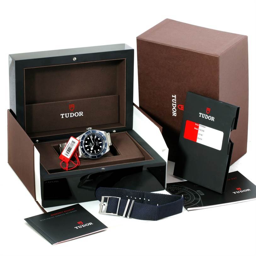 The image shows a Tudor Heritage Black Bay 79220 Men's Stainless Steel Black Dial 79220 Men's Stainless Steel Black Dial watch angled in its box with additional accessories and documentation around it.