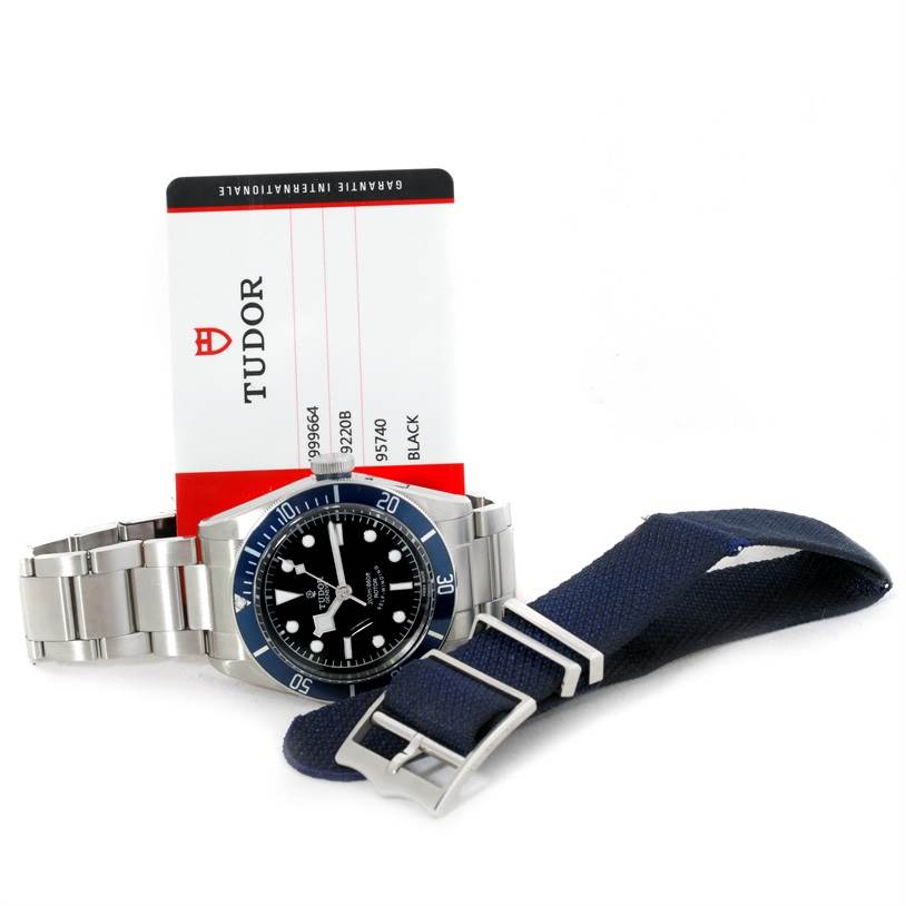 The Tudor Heritage Black Bay 79220 Men's Stainless Steel Black Dial 79220 Men's Stainless Steel Black Dial watch is shown at an angle with a metal bracelet, warranty card, and additional blue fabric strap.