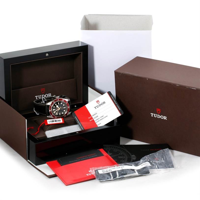 The image shows a Tudor Heritage Black Bay 79220R Men
s Stainless Steel Black Dial 79220R Men
s Stainless Steel Black Dial watch in its packaging, including the watch box, manuals, warranty card, and extra strap.