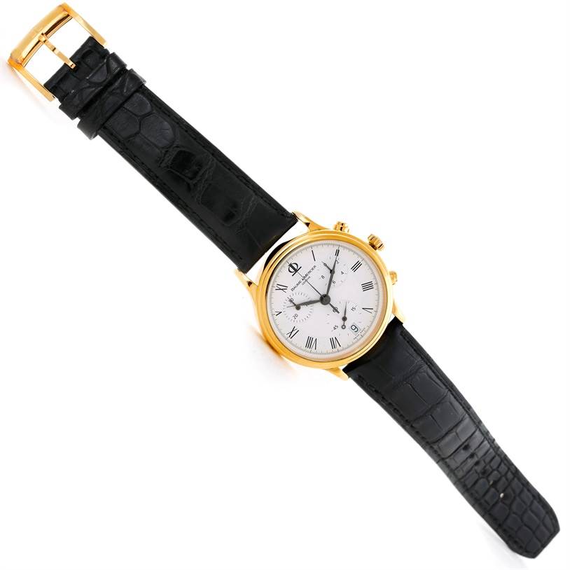 The image shows a Baume & Mercier Classima Men's Yellow Gold White Dial Men's Yellow Gold White Dial watch angled diagonally, displaying its face, black leather strap, and gold case.
