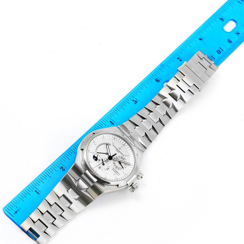 The Vacheron Constantin Overseas 47450/B01A-9226 Men's Stainless Steel Silver Dial 47450/B01A-9226 Men's Stainless Steel Silver Dial watch is shown from a top-down angle, with the full watch and bracelet laid out beside a blue ruler.