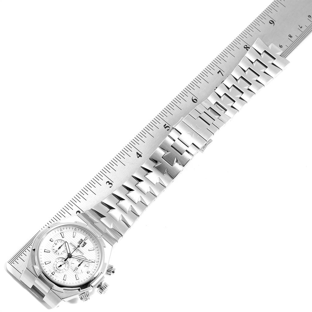 The image shows a top-down view of the Vacheron Constantin Overseas 49150 Men's Stainless Steel Silver Dial 49150 Men's Stainless Steel Silver Dial watch and its strap alongside a ruler.