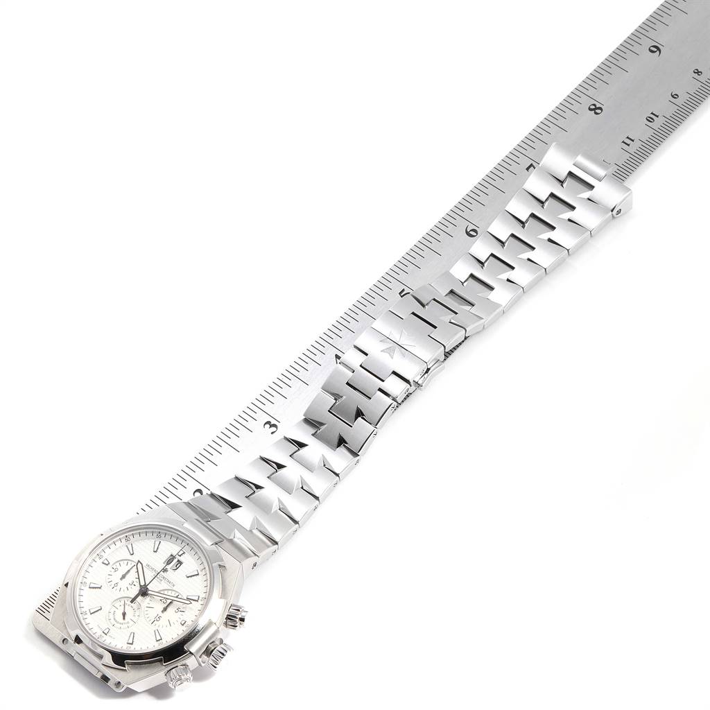 The Vacheron Constantin Overseas 49150 Men's Stainless Steel Silver Dial 49150 Men's Stainless Steel Silver Dial watch is shown from a side angle, displaying the dial and metal strap on a ruler for measurement.