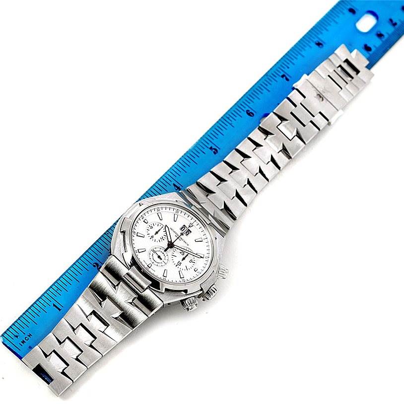 The image shows the Vacheron Constantin Overseas 49150 Men's Stainless Steel Silver Dial 49150 Men's Stainless Steel Silver Dial watch laid flat with a metal bracelet next to a blue ruler for measurement.