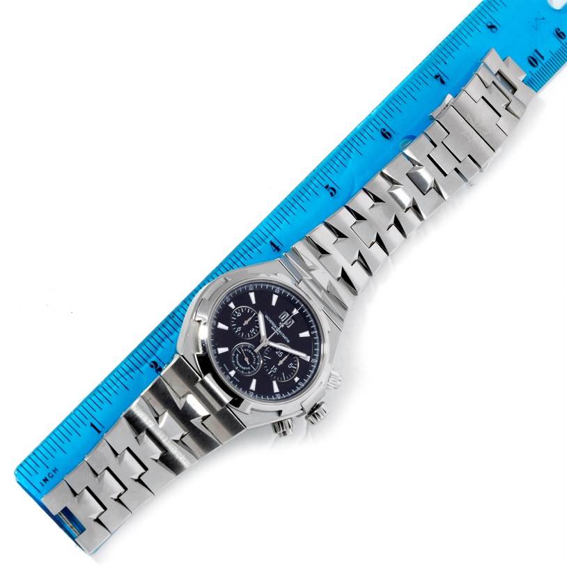 The image shows a top-down view of the Vacheron Constantin Overseas 49150.B01A.9097 Men's Stainless Steel Black Dial 49150.B01A.9097 Men's Stainless Steel Black Dial watch and its metal bracelet, aligned with a blue ruler.