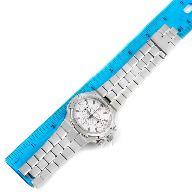 The image shows a Vacheron Constantin Overseas 49140/423A-8790 Men's Stainless Steel Silver Dial 49140/423A-8790 Men's Stainless Steel Silver Dial model watch laid flat next to a blue ruler, displaying its face, bracelet, and pushers.