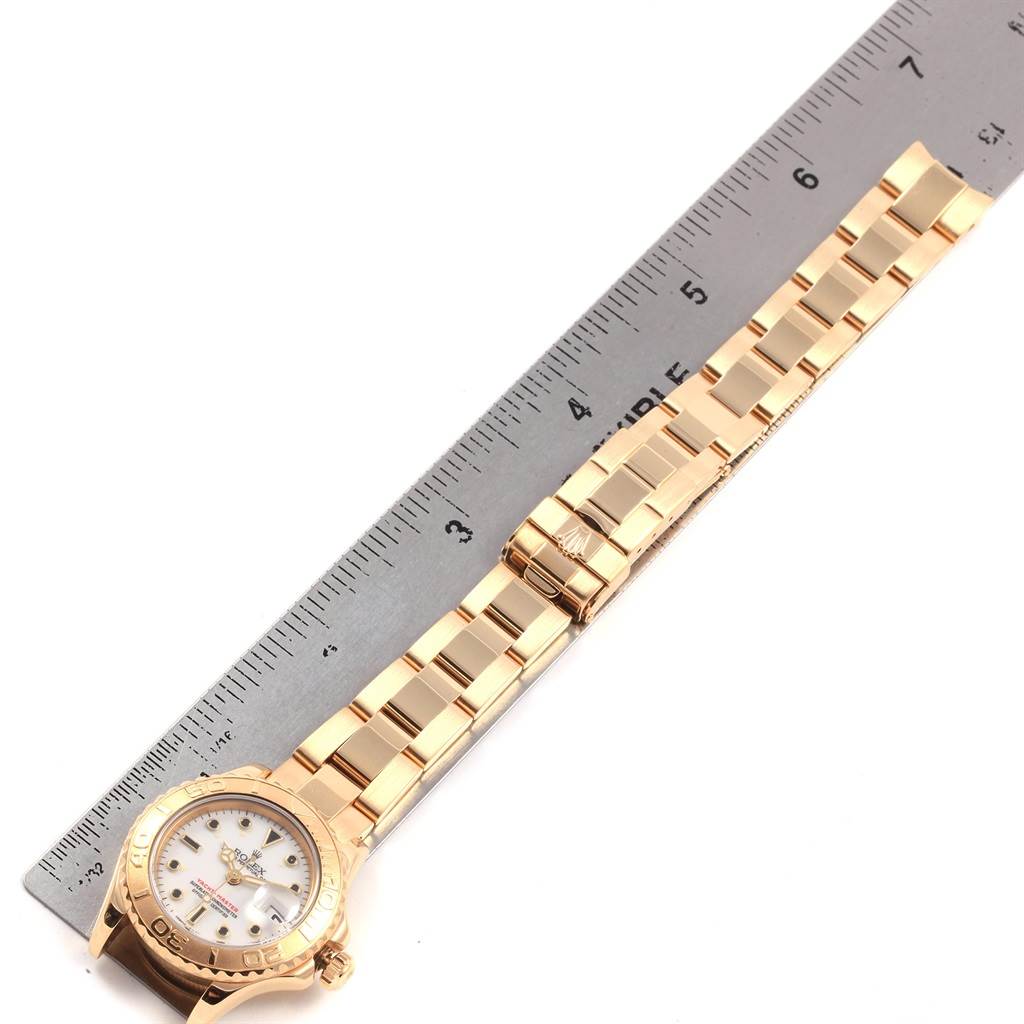 This image shows a Rolex Yacht-Master 69628 Women's Yellow Gold White Dial 69628 Women's Yellow Gold White Dial watch with a gold bracelet, laid flat next to a ruler for size reference.