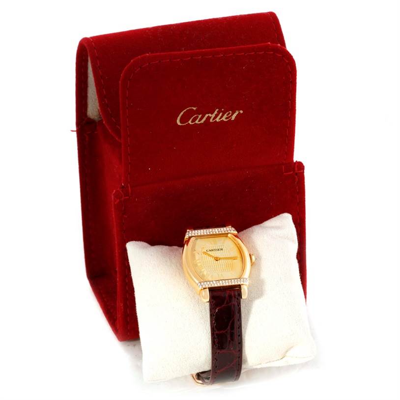The image shows a Cartier Tortue Women's Yellow Gold Champagne Dial Women's Yellow Gold Champagne Dial watch with a gold case and dark strap, placed on a cushion in front of a red Cartier box.