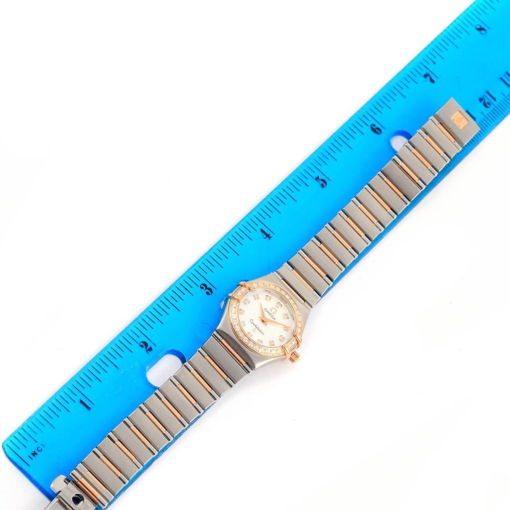 This image shows an Omega 1360.75.00 Women's Steel and Gold (two tone) Mother of Pearl Dial 1360.75.00 Women's Steel and Gold (two tone) Mother of Pearl Dial Constellation My Choice Mini Diamond Watch 1360.75.00 from a top-down angle beside a blue ruler for scale.