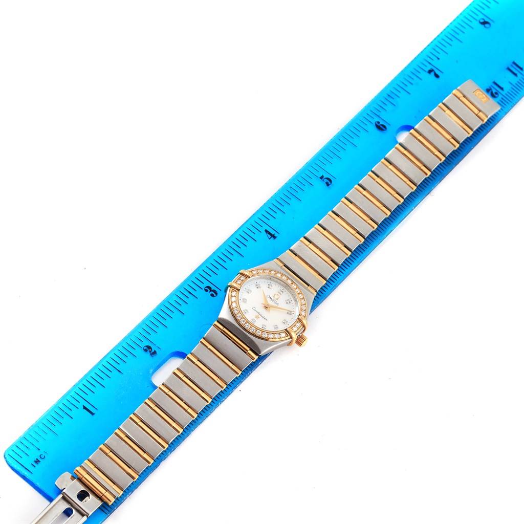 The image shows an overhead view of the Omega 1267.75.00 Women's Steel and Gold (two tone) Mother of Pearl Dial 1267.75.00 Women's Steel and Gold (two tone) Mother of Pearl Dial Constellation Mini Mother of Pearl Diamond Watch 1267.75.00 beside a blue ruler.