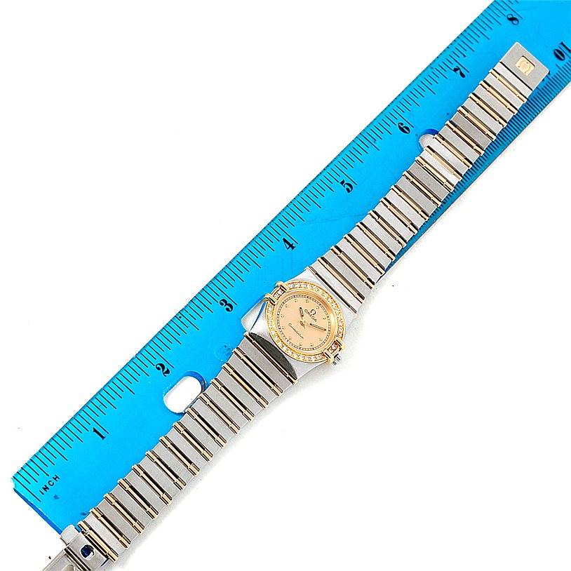 The image shows the Omega Women
s Steel and Gold (two tone) Champagne Dial Women
s Steel and Gold (two tone) Champagne Dial Constellation Mini Steel 18K Yellow Gold Diamond Watch model laid flat next to a blue ruler.