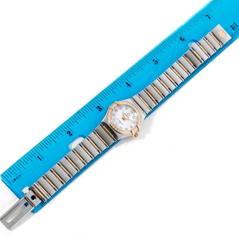 The image shows an Omega 1360.75.00 Women
s Steel and Gold (two tone) Mother of Pearl Dial 1360.75.00 Women
s Steel and Gold (two tone) Mother of Pearl Dial Constellation My Choice Mini Diamond Watch 1360.75.00 laid flat beside a blue ruler.