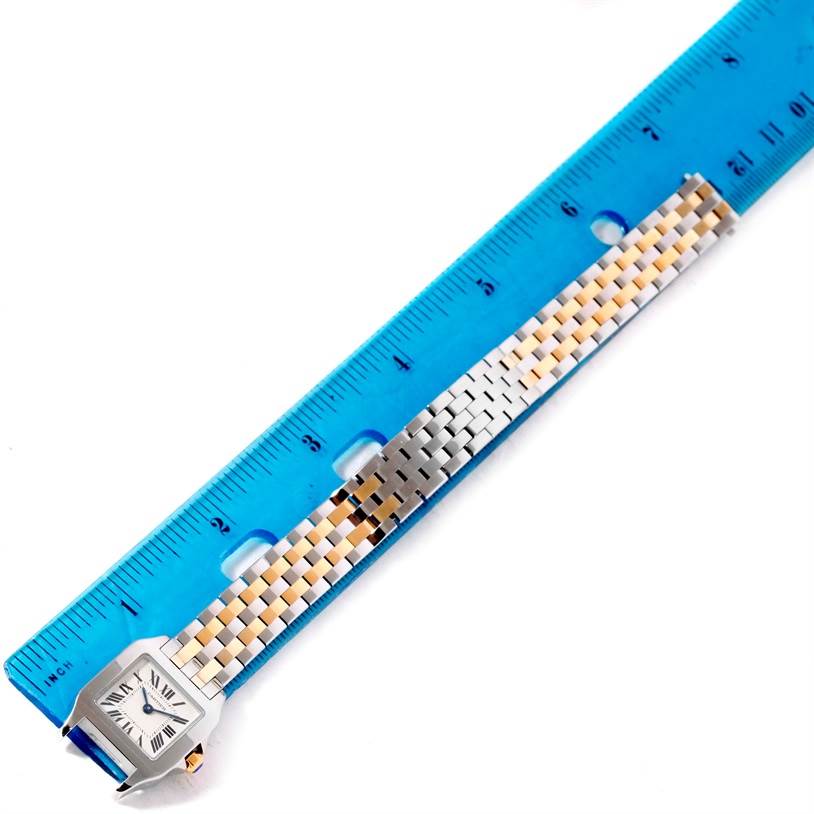 The image shows a Santos Demoiselle watch by Cartier laid next to a blue ruler, displaying its face and metal bracelet.
