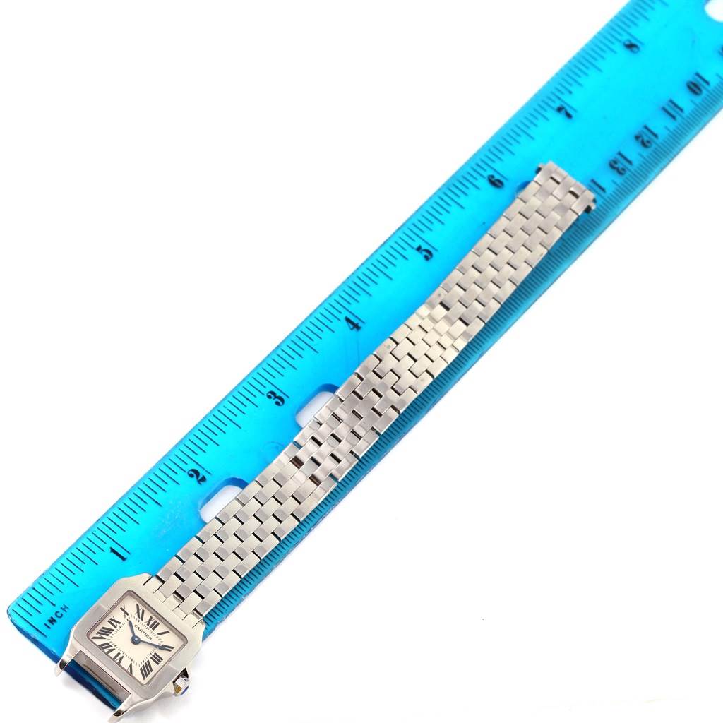 The Santos Demoiselle watch by Cartier is shown laying flat next to a blue ruler, displaying its face and metal bracelet.