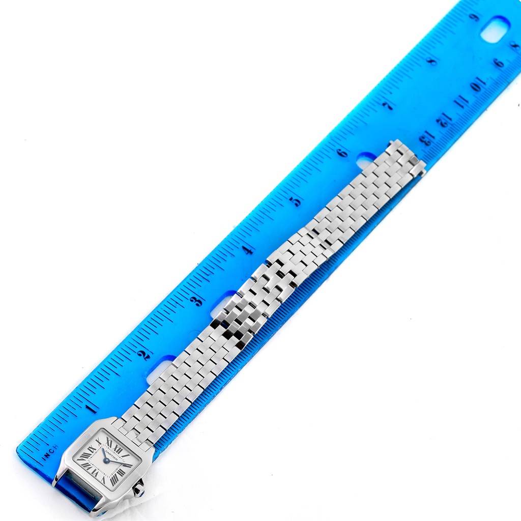 The image shows a Santos Demoiselle model by Cartier placed next to a blue ruler, displaying the watch face and metal bracelet.