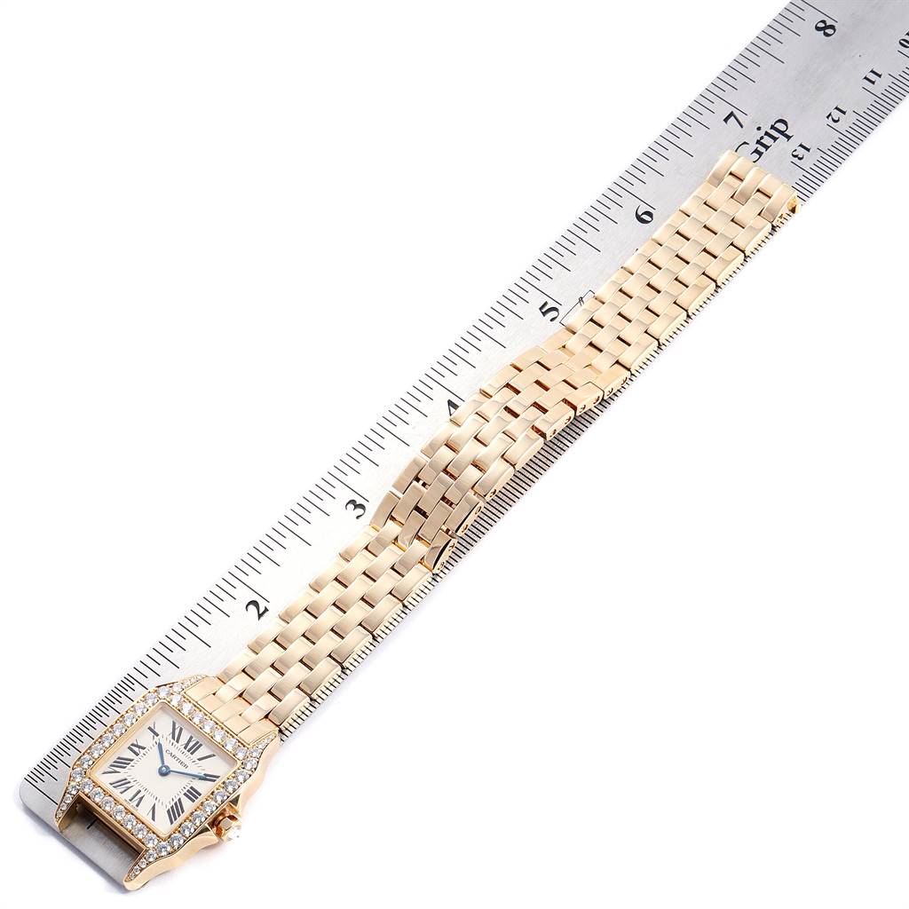 The image shows a Cartier Santos Demoiselle WF9002Y7 Women
s Yellow Gold Silver Dial WF9002Y7 Women
s Yellow Gold Silver Dial model watch laying flat beside a ruler, displaying its face and full bracelet.
