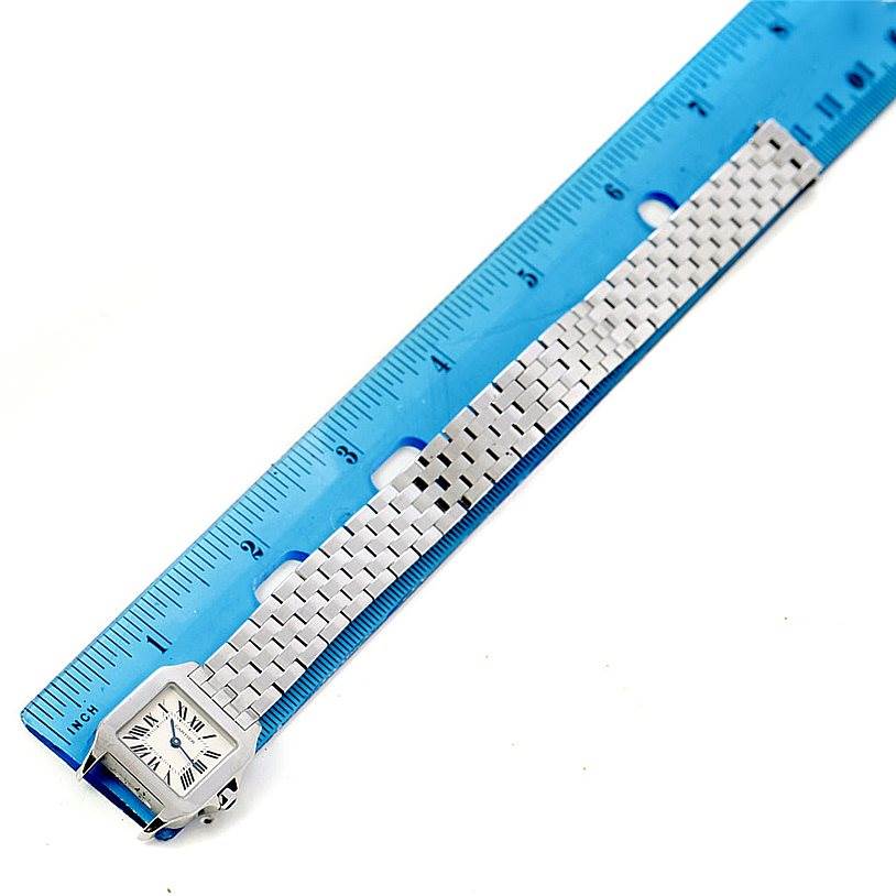 The image shows a Cartier Santos Demoiselle W25064Z5 Women
s Stainless Steel Silver Dial W25064Z5 Women
s Stainless Steel Silver Dial watch placed alongside a blue ruler, capturing the face and the metal bracelet.