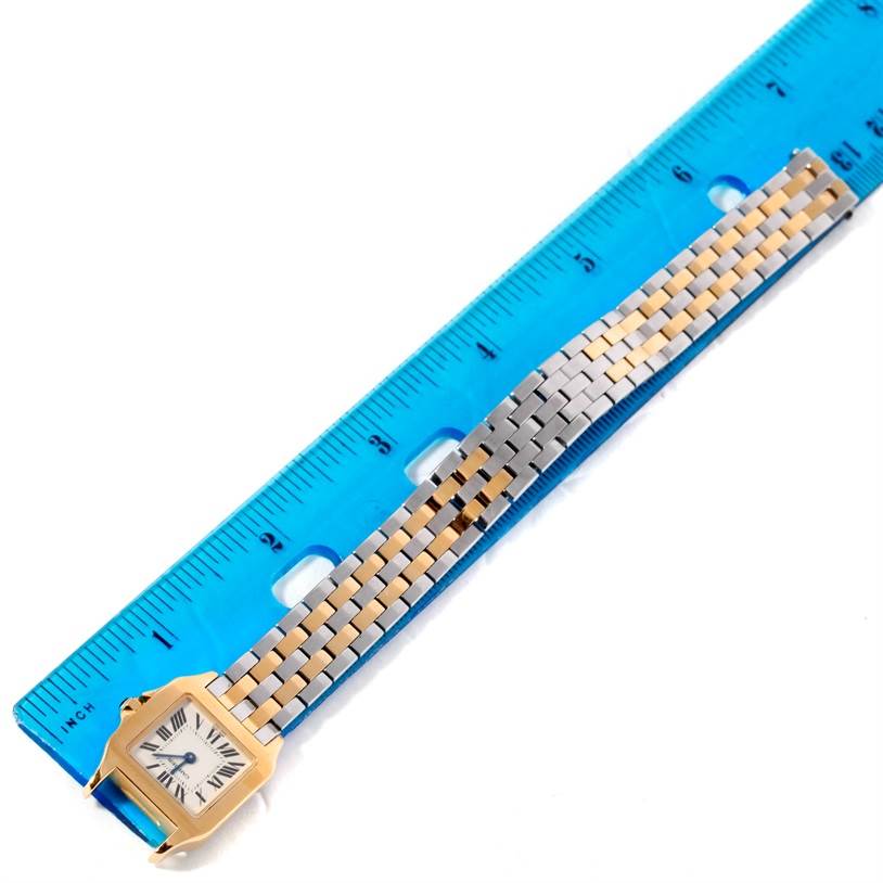 The image shows a Cartier Santos Demoiselle W25063Z6 Women's Yellow Gold Silver Dial W25063Z6 Women's Yellow Gold Silver Dial watch laid flat next to a blue ruler, displaying the face and metal band.