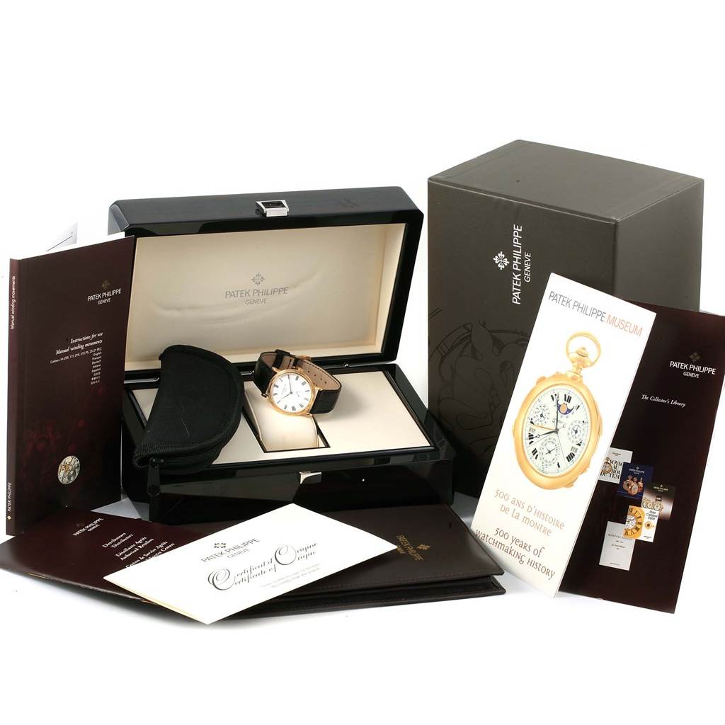 This image shows a Patek Philippe Calatrava 5119J Men's Yellow Gold White Dial 5119J Men's Yellow Gold White Dial watch in its open box, along with booklets and documentation placed around it.