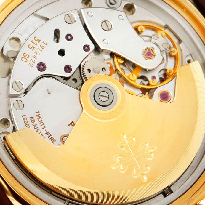 The image shows an angled close-up of the movement of a Patek Philippe Calatrava 5012 Men
s Yellow Gold Beige Dial 5012 Men
s Yellow Gold Beige Dial watch, highlighting its intricate mechanism and rotor.