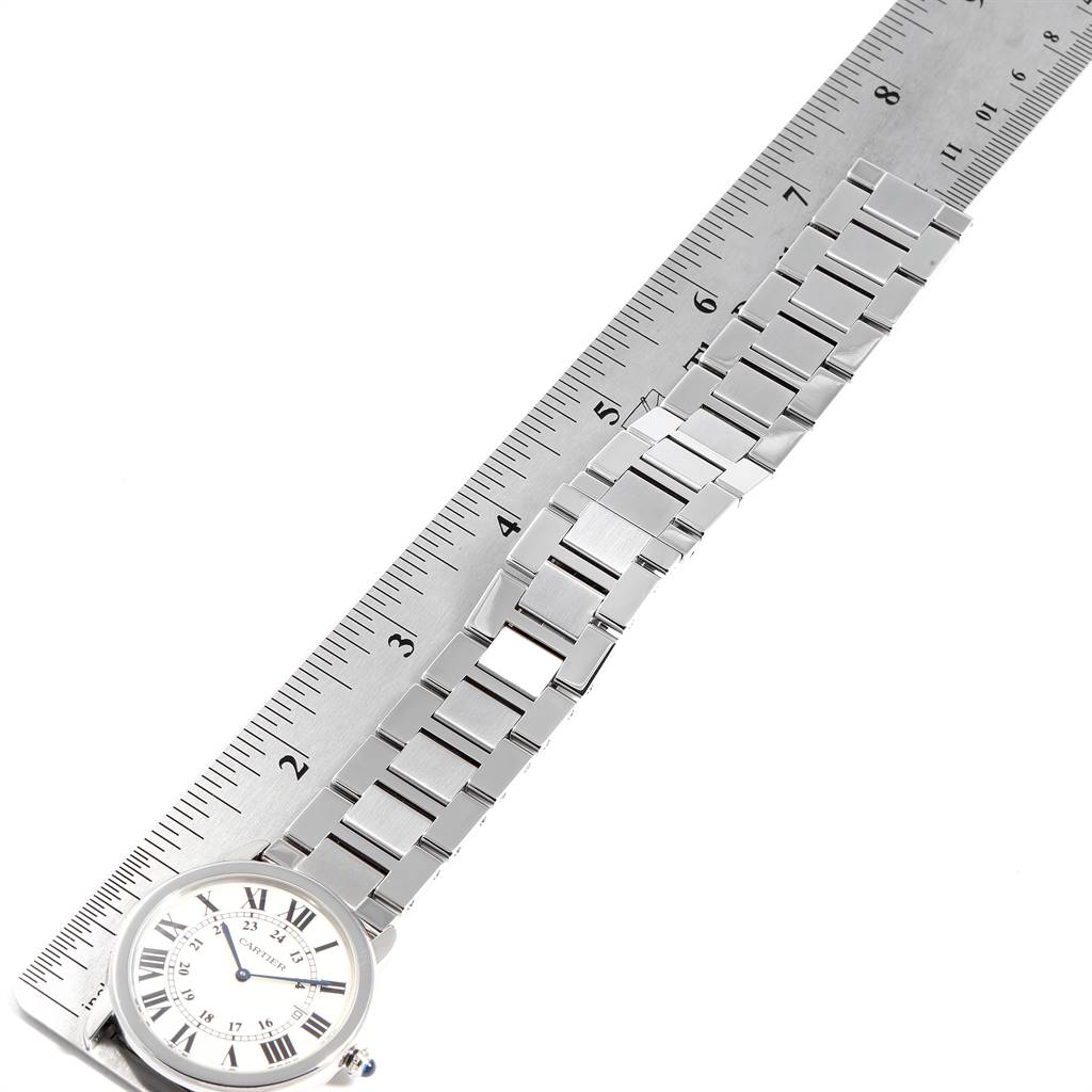 The image shows the Ronde model by Cartier, featuring a detailed view of the watch face and the full length of the metal bracelet beside a ruler.