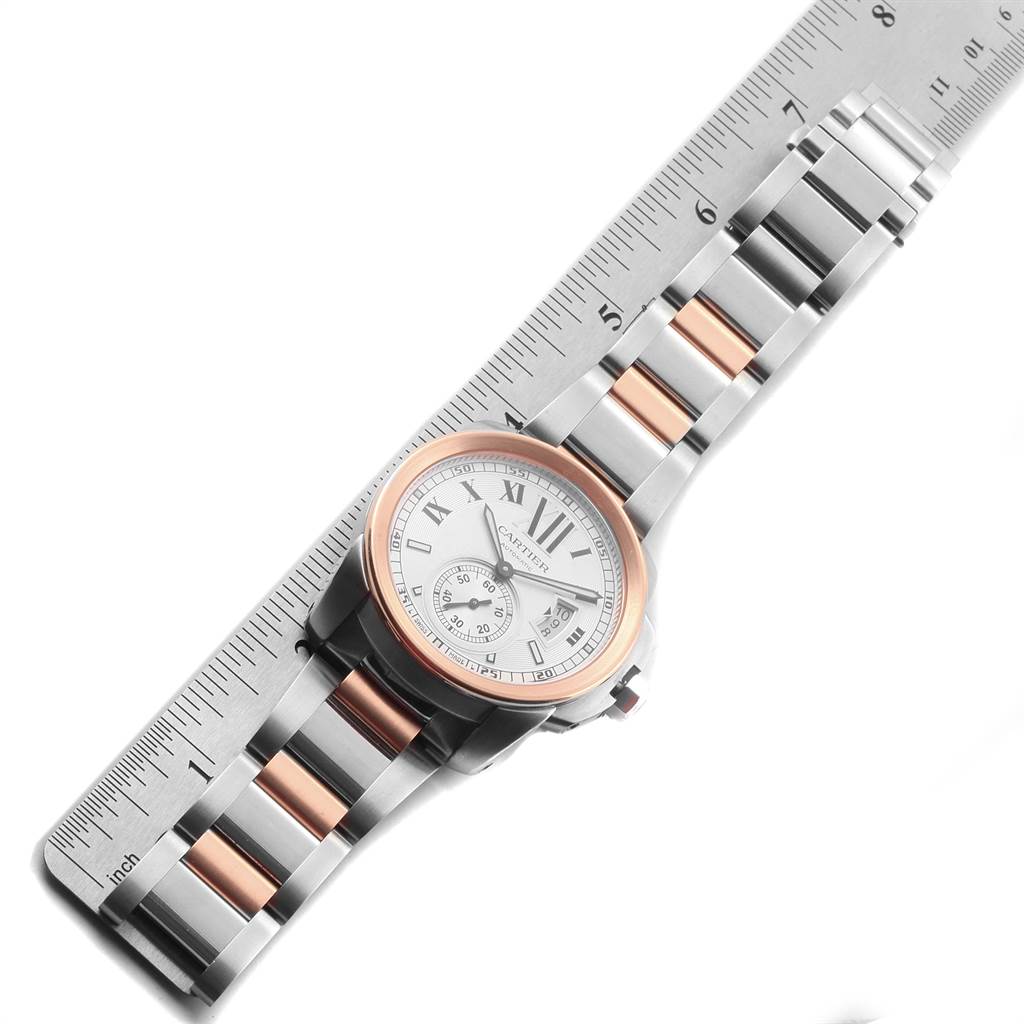 The image shows a Calibre de Cartier watch by Cartier placed diagonally alongside a ruler, highlighting its bracelet and face.