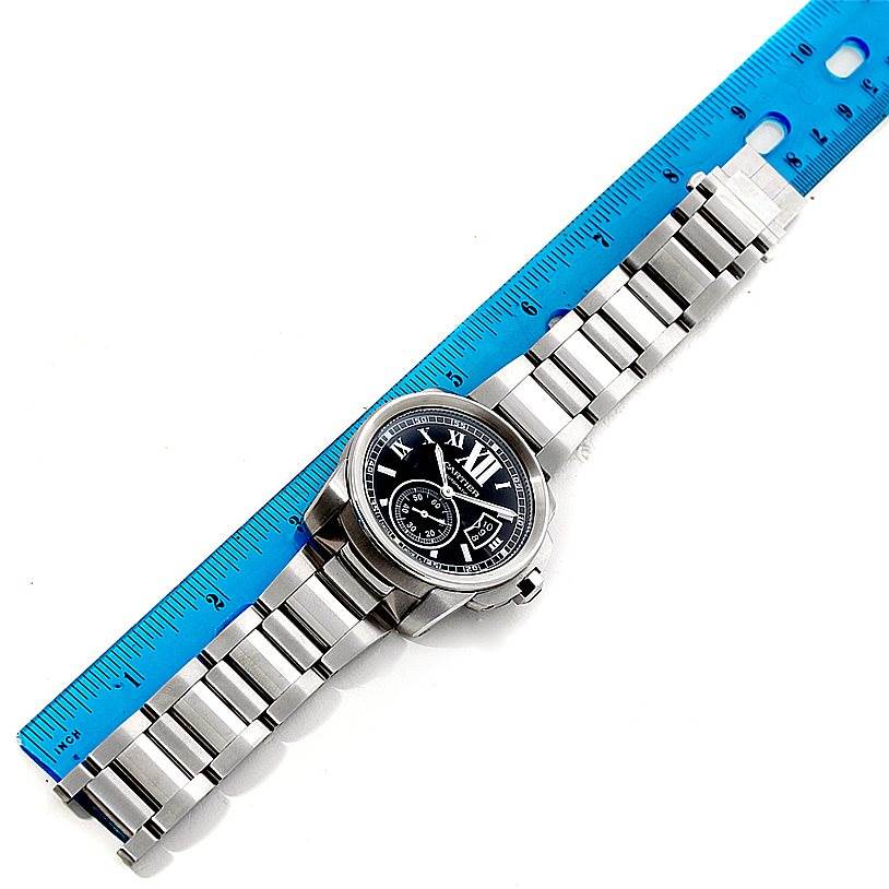 The image shows a top view of a Calibre de Cartier watch by Cartier, including the face and metal bracelet next to a blue ruler.