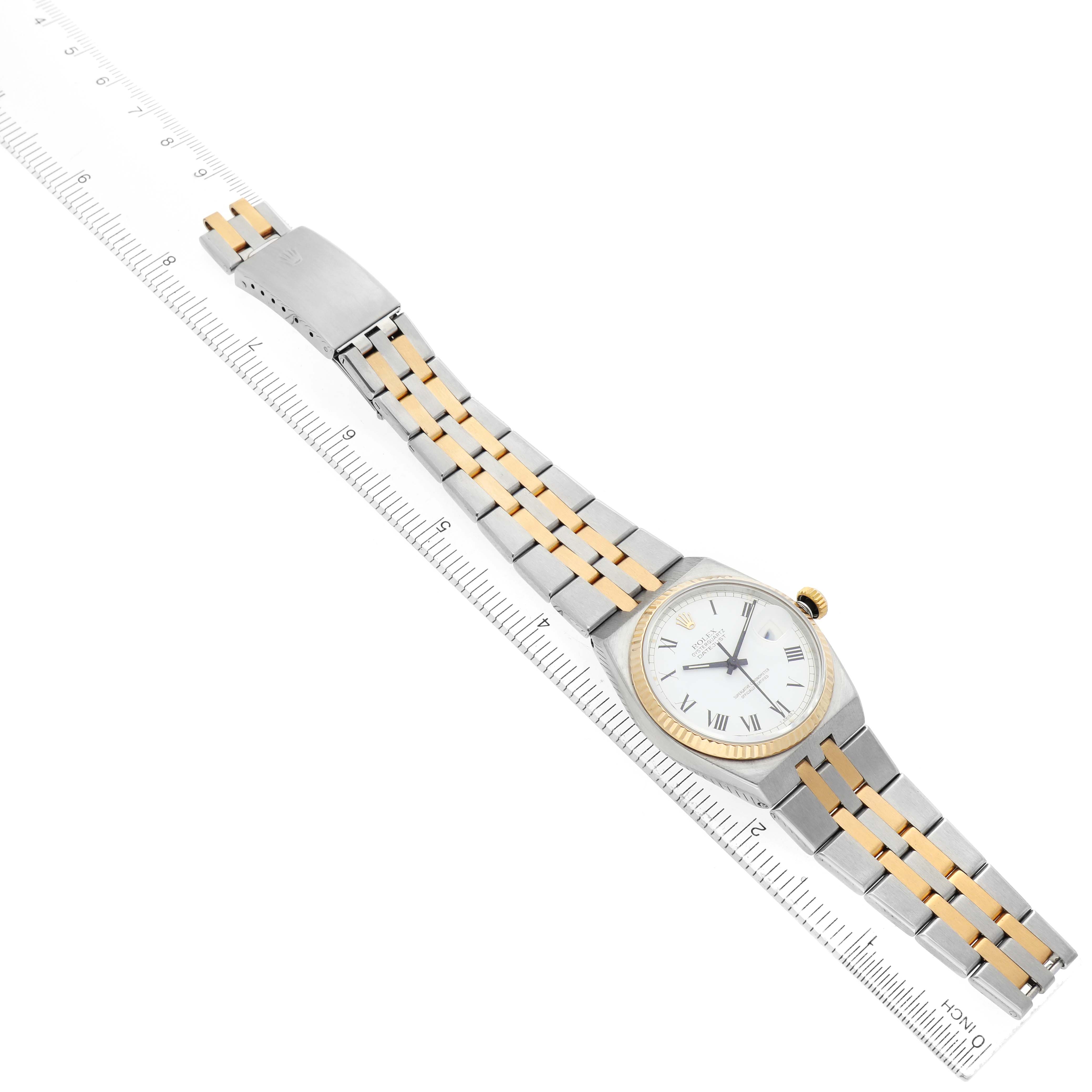 The Rolex Oysterquartz 17013 Men's Steel and Gold (two tone) White Dial watch is shown at a diagonal angle, displaying its bezel, dial, bracelet, and clasp on a ruler background.