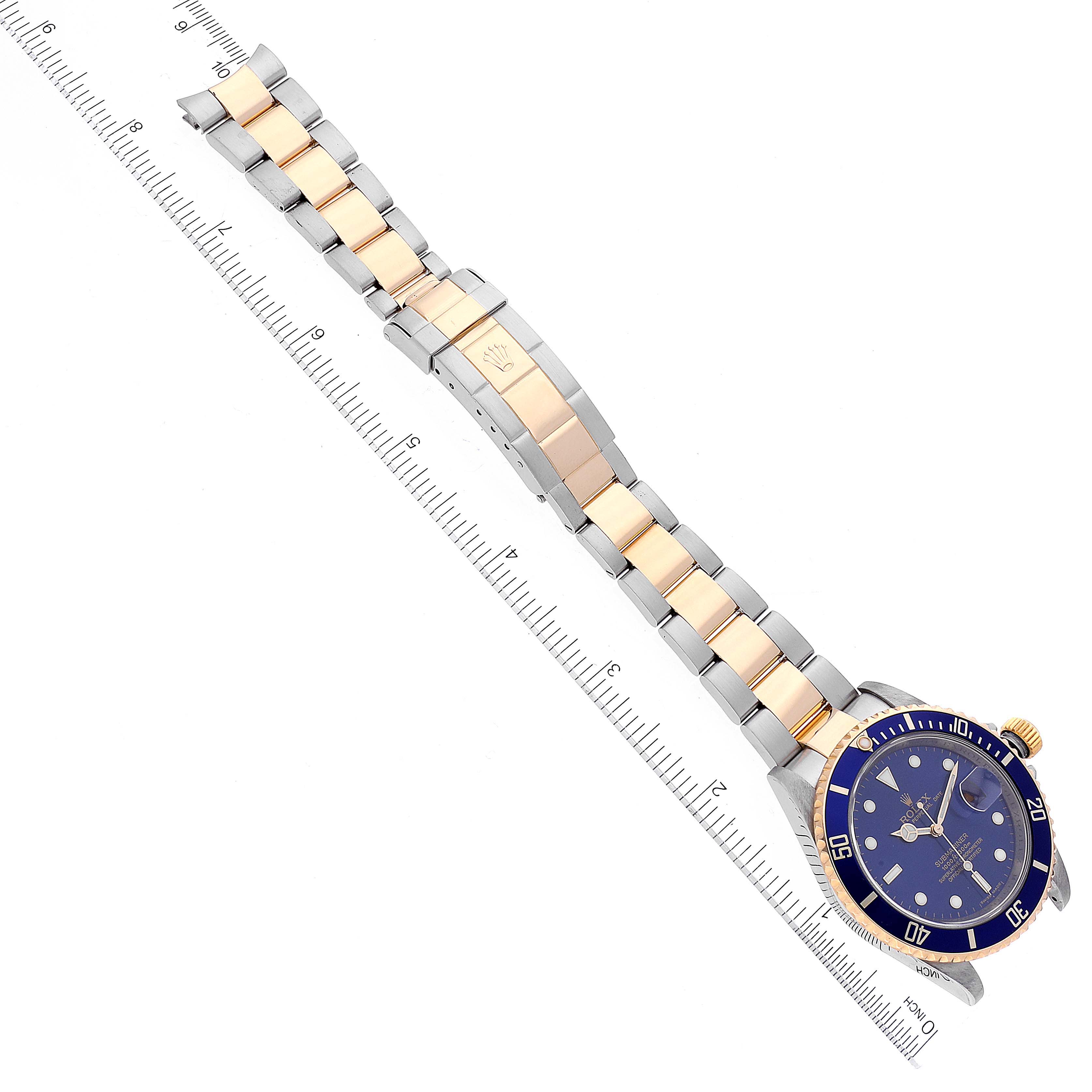 The image shows a top-down view of a Rolex Submariner 16613 Men's Steel and Gold (two tone) Blue Dial 16613 Men's Steel and Gold (two tone) Blue Dial watch, featuring the entire bracelet, clasp, and face, positioned alongside a ruler.