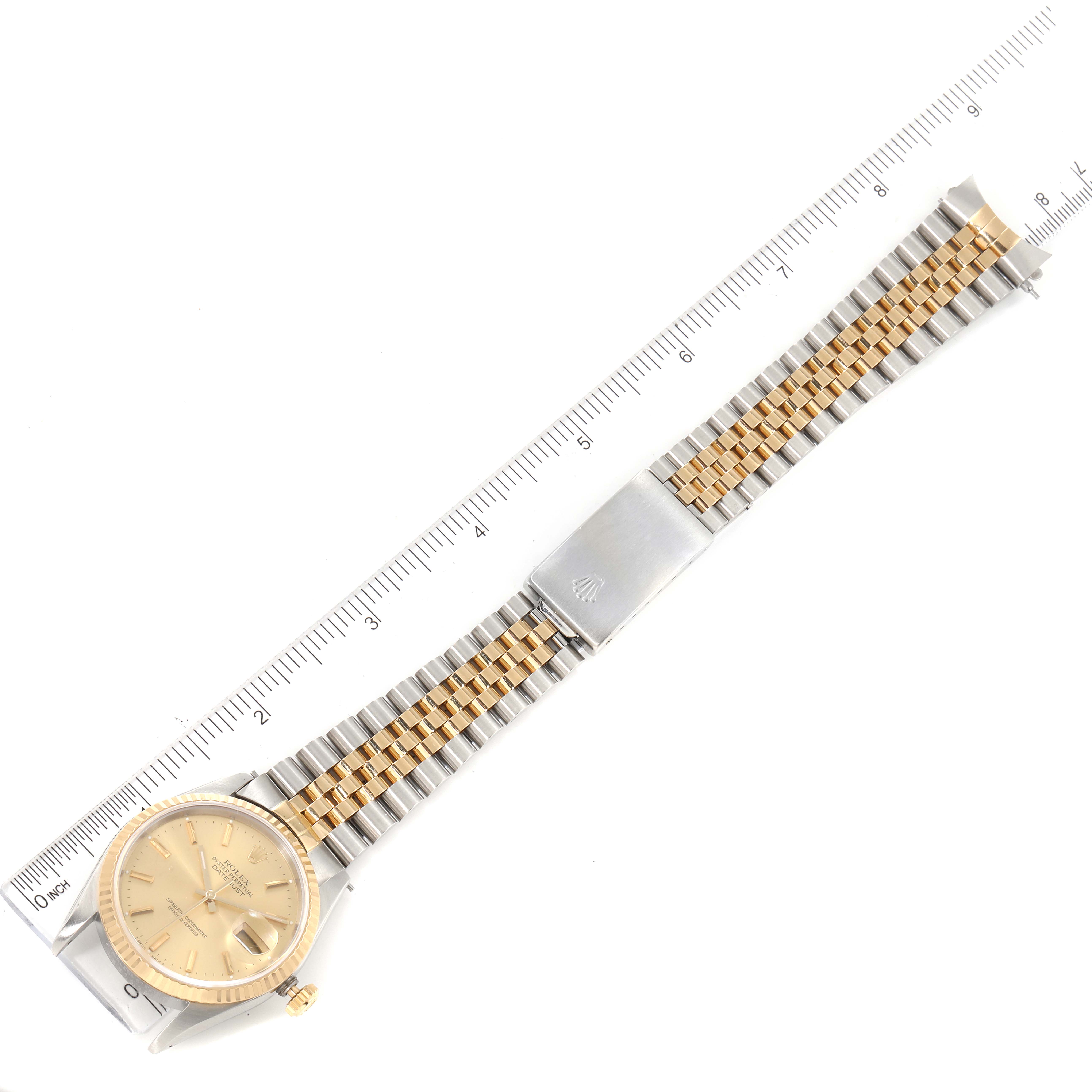 The Rolex Datejust 16233 Men
s Steel and Gold (two tone) Champagne Dial 16233 Men
s Steel and Gold (two tone) Champagne Dial watch is shown in a top-down angle, displaying the face, bracelet, and clasp next to a ruler.