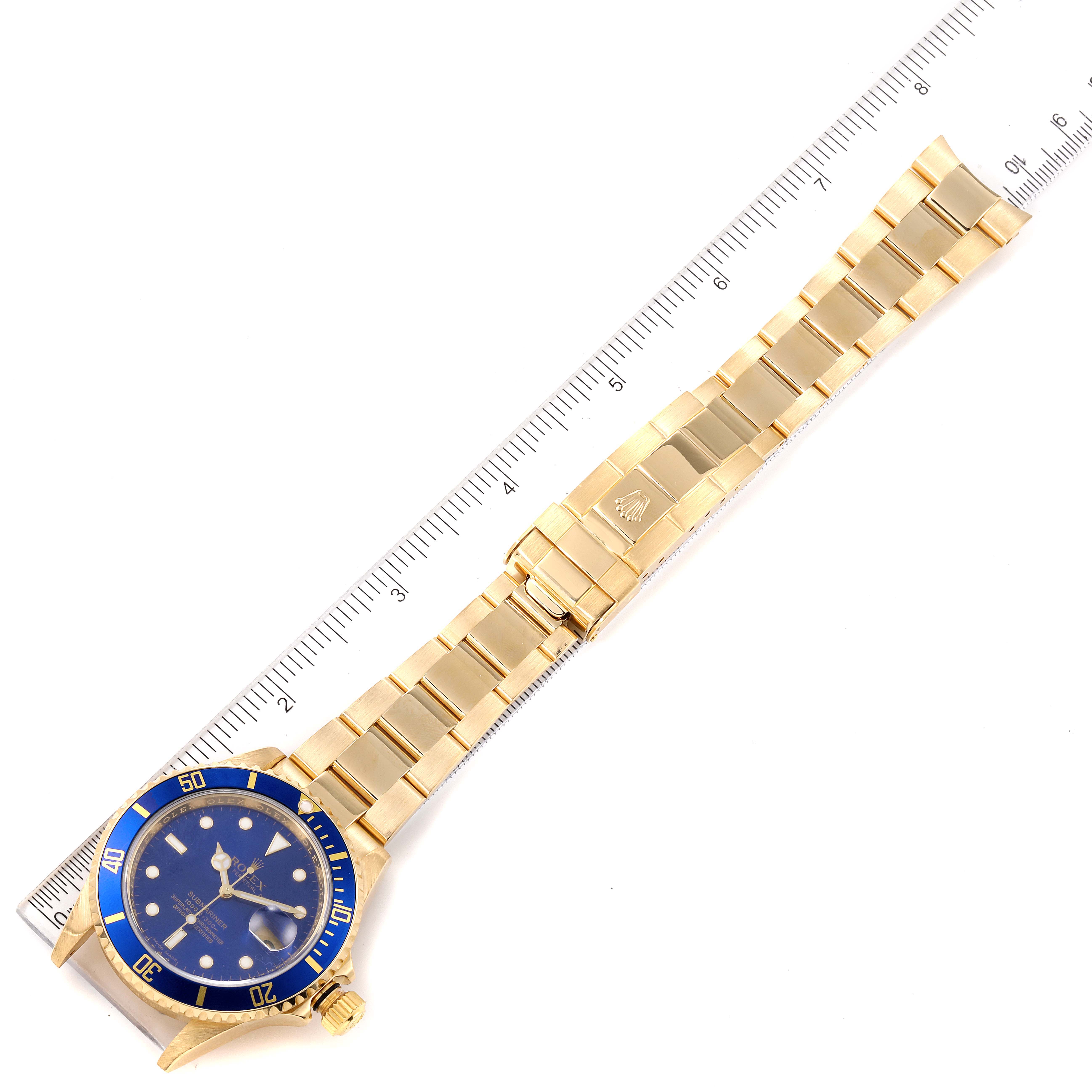 A Rolex Submariner 16618 Men's Yellow Gold Blue Dial 16618 Men's Yellow Gold Blue Dial watch is shown laying flat, displaying the face, bezel, bracelet, and clasp against a background with a ruler.