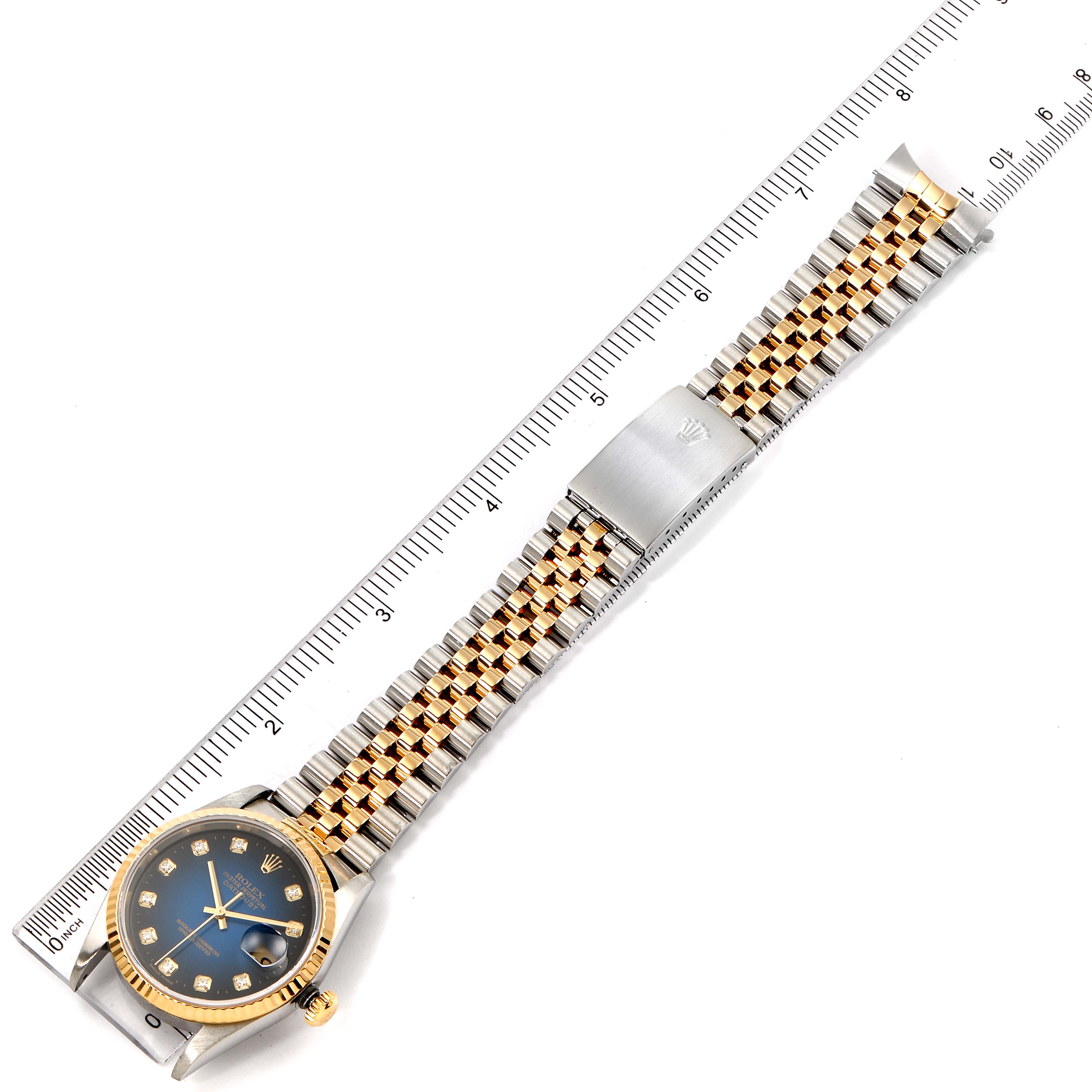 The Rolex Datejust 16233 Men's Steel and Gold (two tone) Blue Dial 16233 Men's Steel and Gold (two tone) Blue Dial watch is shown from a top-down angle, highlighting the dial, bezel, bracelet, and clasp beside a ruler.