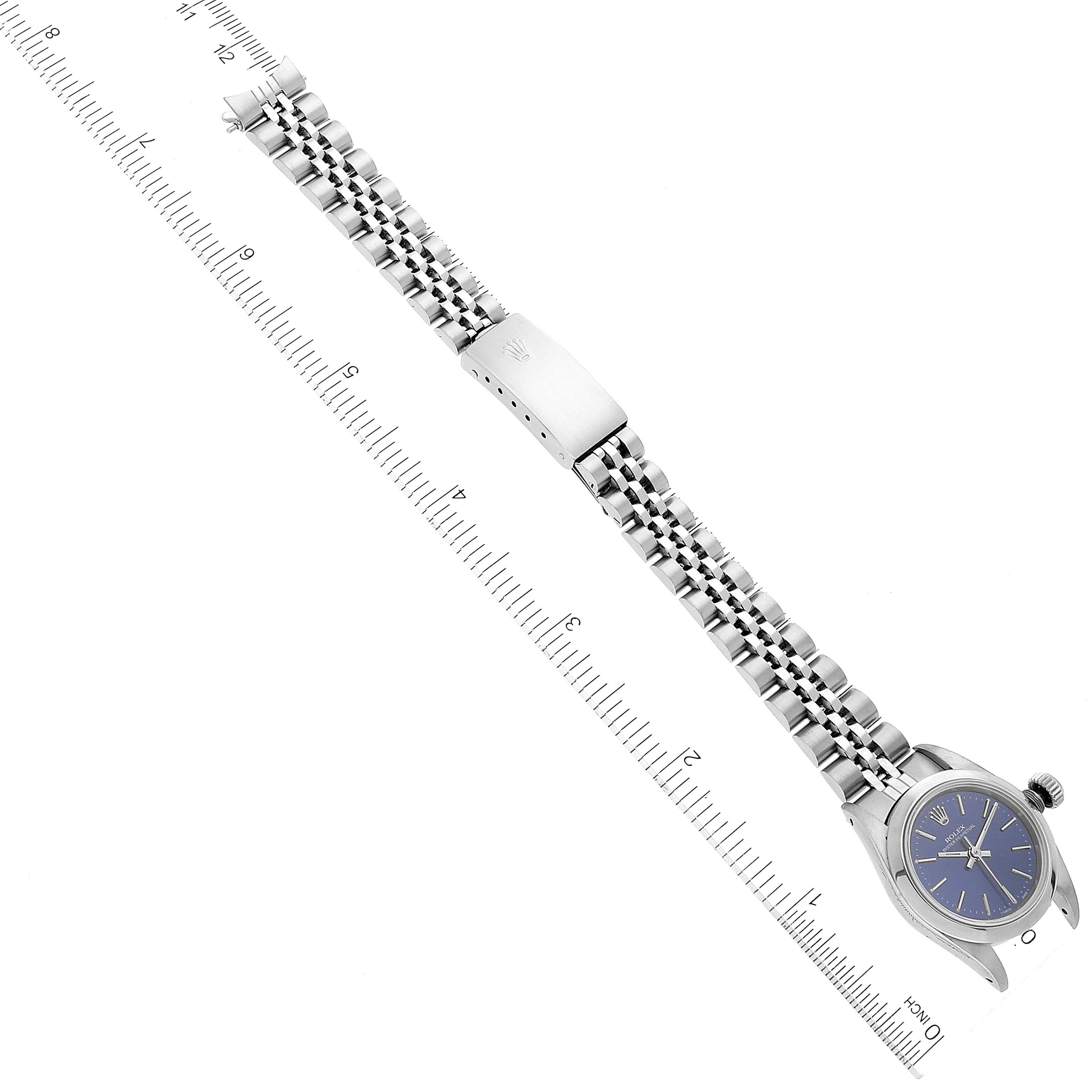 The image shows a Rolex Oyster Perpetual 67180 Women
s Stainless Steel Blue Dial 67180 Women
s Stainless Steel Blue Dial watch laid out flat with its metal bracelet and blue dial, including a ruler for scale.