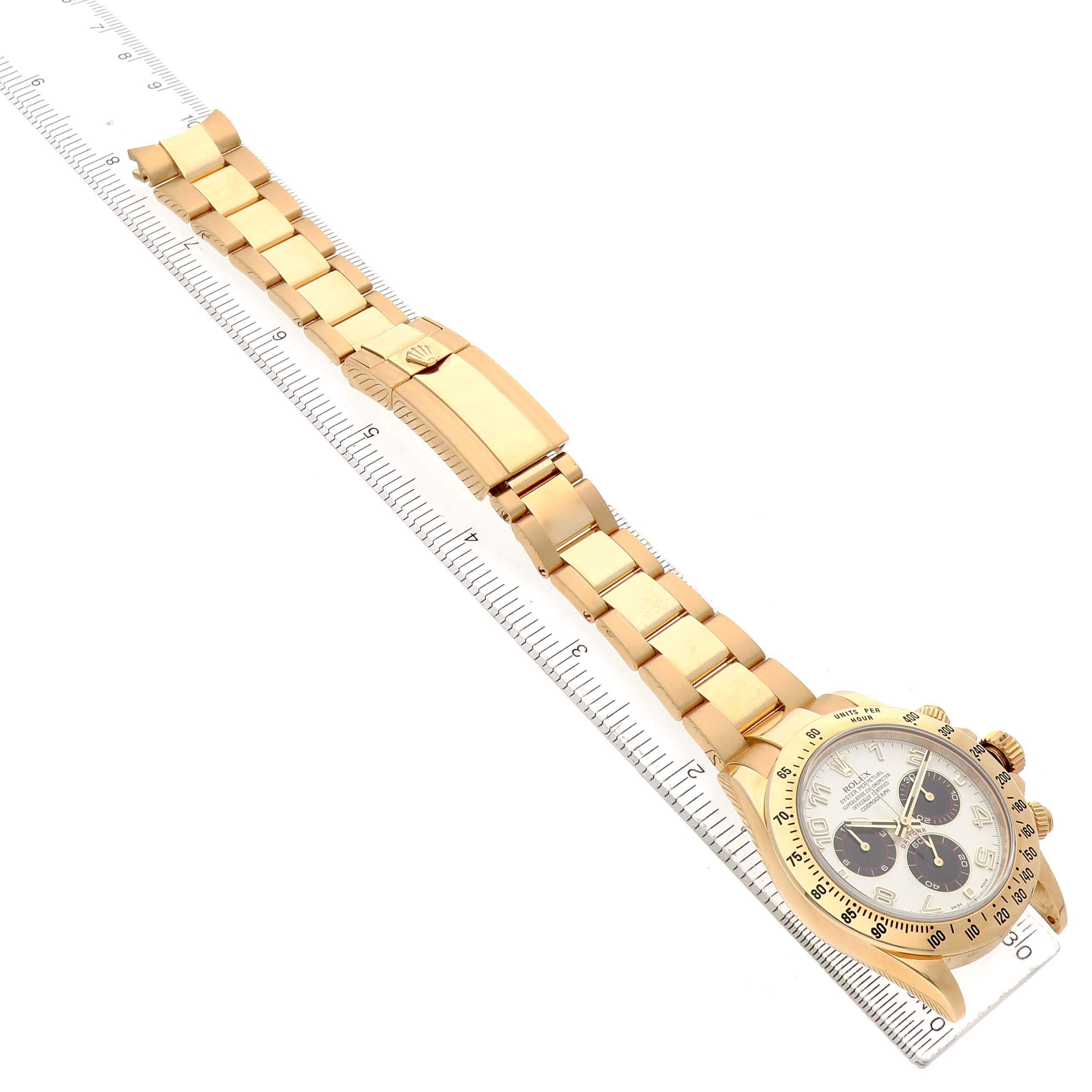 The Rolex Daytona 116528 Men's Yellow Gold White Dial is shown at an angle highlighting the bracelet, clasp, and watch face placed alongside a ruler.