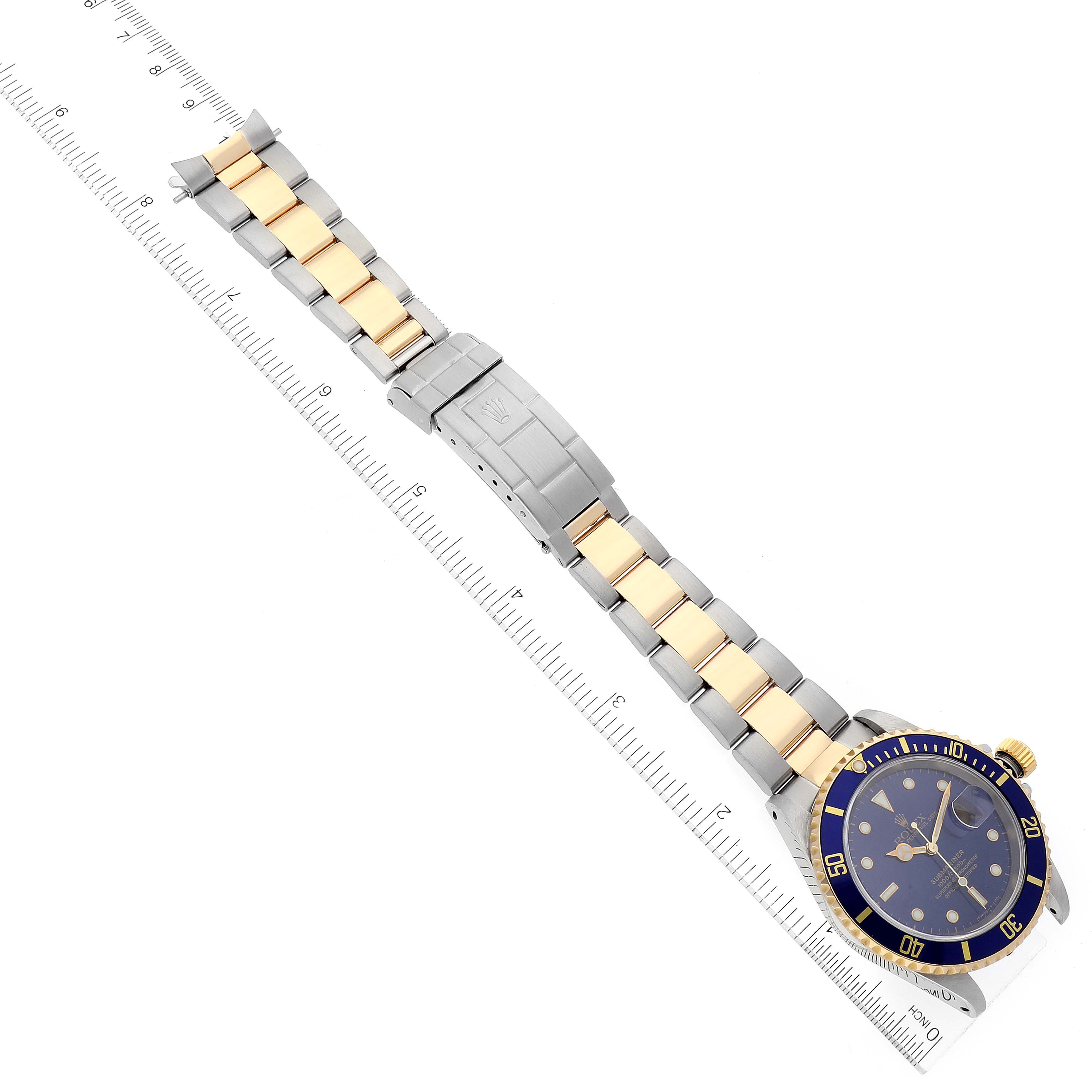 The image shows the Rolex Submariner 16613 Men's Steel and Gold (two tone) Blue Dial 16613 Men's Steel and Gold (two tone) Blue Dial watch laid flat, highlighting the face, bezel, bracelet, and clasp next to a ruler.
