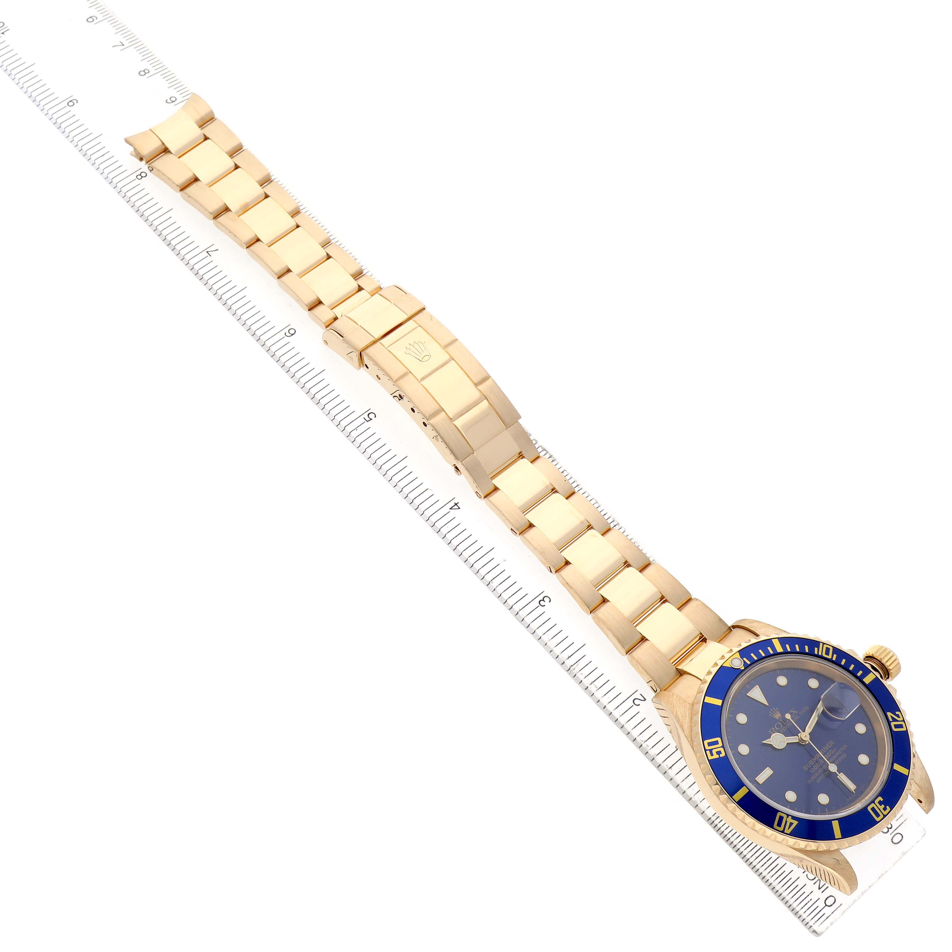 The Rolex Submariner 16618 Men's Yellow Gold Blue Dial watch is shown from an angled top view, displaying its blue bezel, dial, and gold bracelet, alongside a ruler.