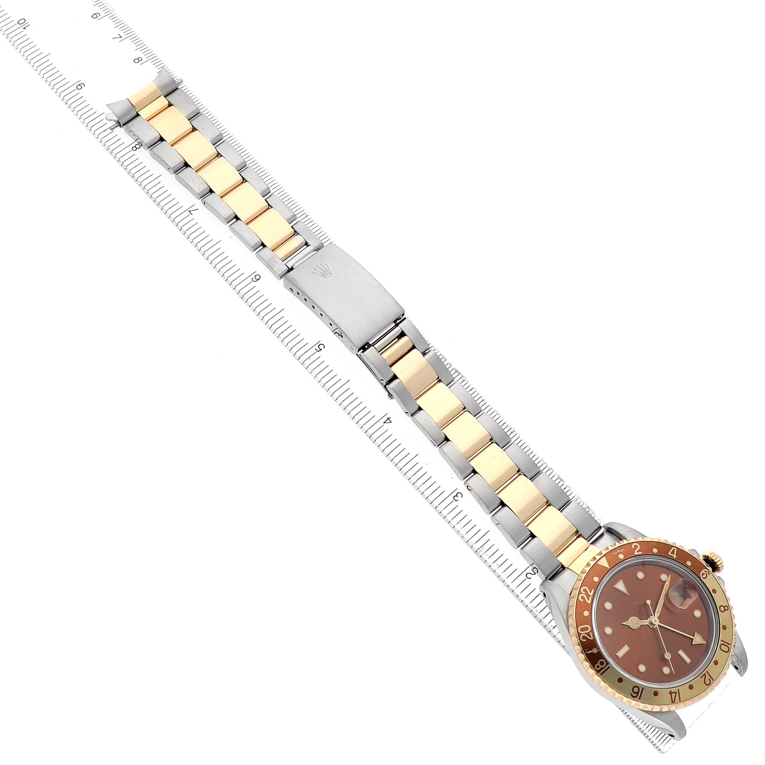 The Rolex GMT-Master 16713 Men's Steel and Gold (two tone) Brown Dial watch is shown from a top-down angle, displaying the face, bezel, and bracelet on a ruler.