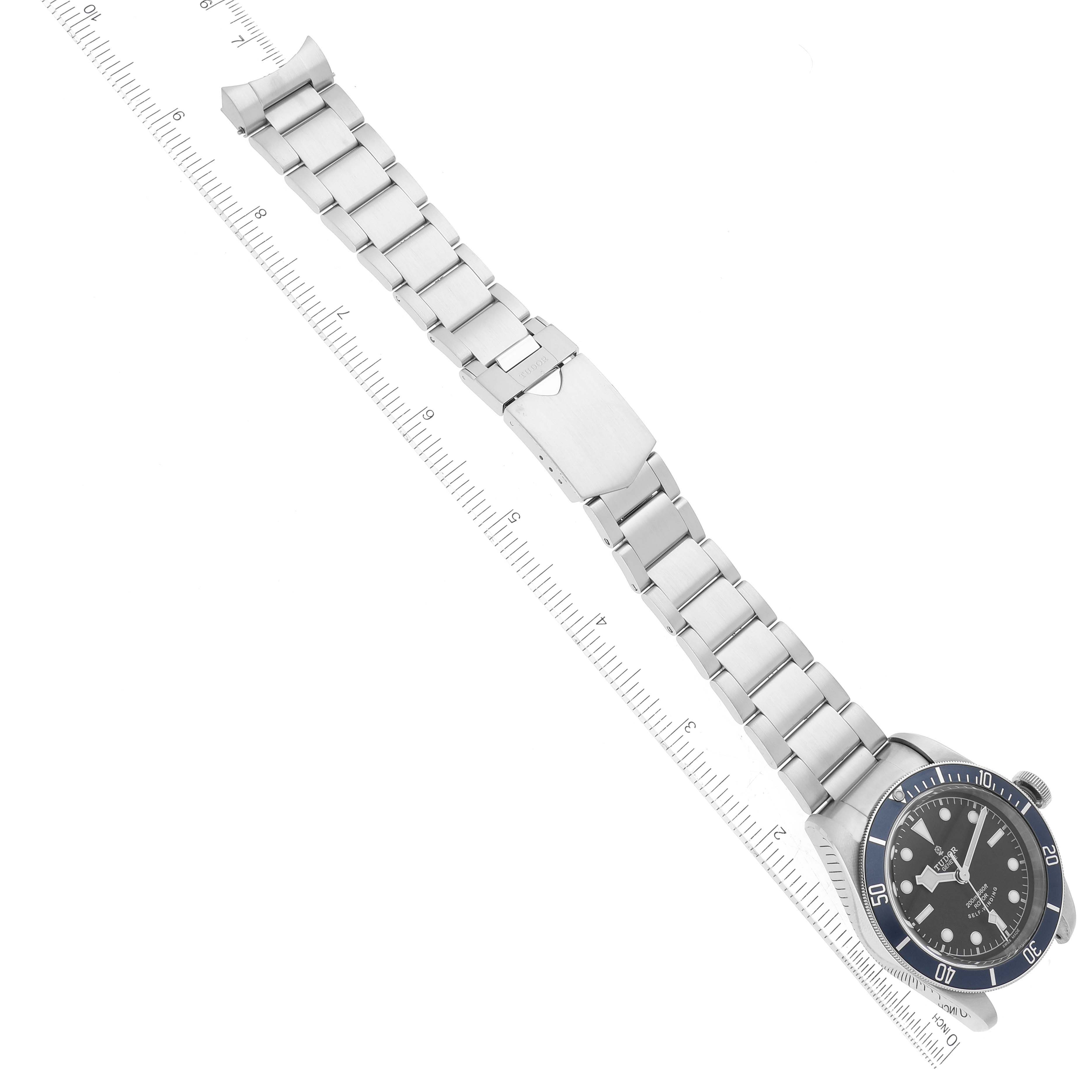 The image shows the Tudor Heritage Black Bay 79220B Men
s Stainless Steel Black Dial 79220B Men
s Stainless Steel Black Dial watch laid out flat with its metal bracelet and clasp visible against a ruler for scale.