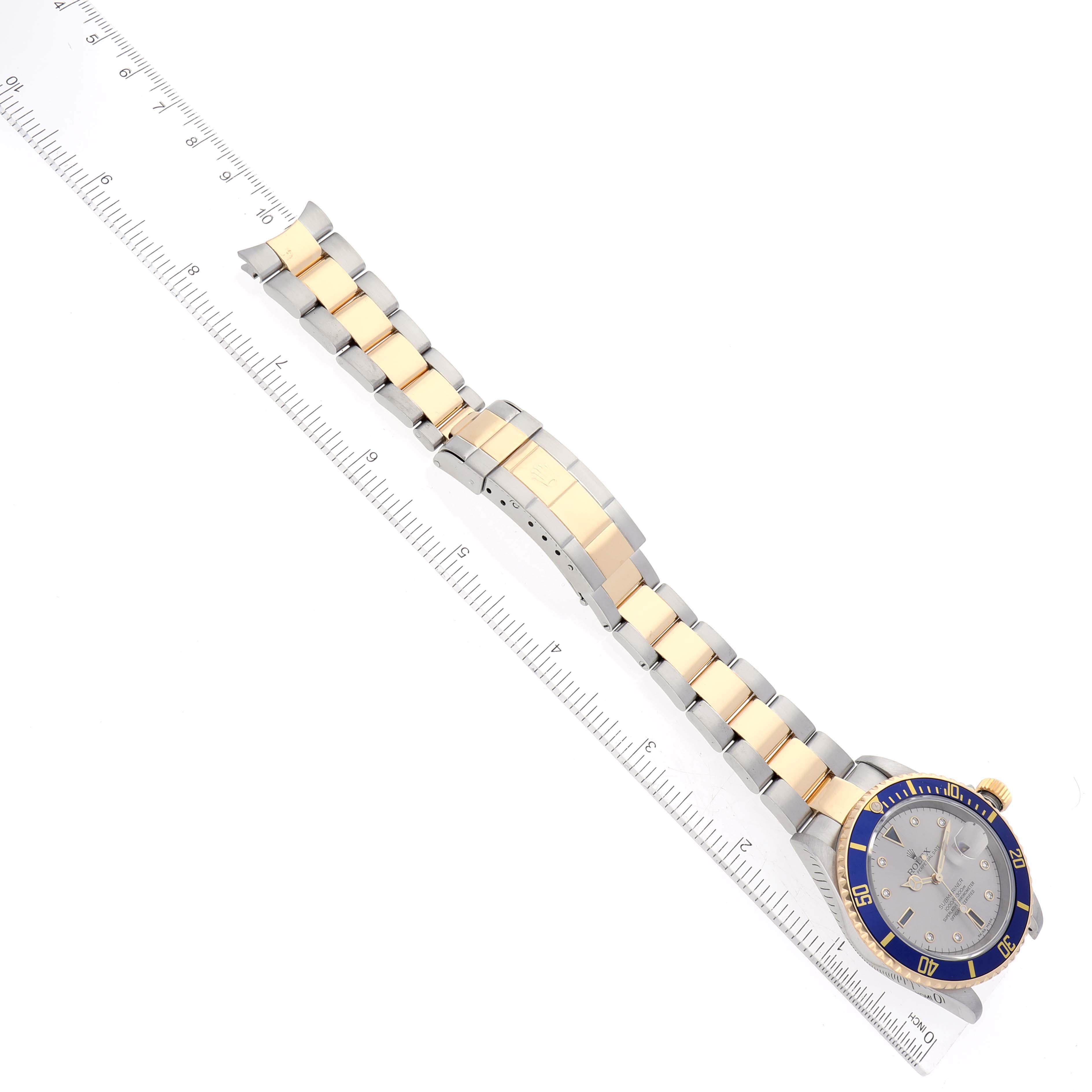 The image shows a Rolex Submariner 16613 Men
s Steel and Gold (two tone) Grey Dial 16613 Men
s Steel and Gold (two tone) Grey Dial watch, highlighting the face, bezel, and metal bracelet, angled alongside a ruler.