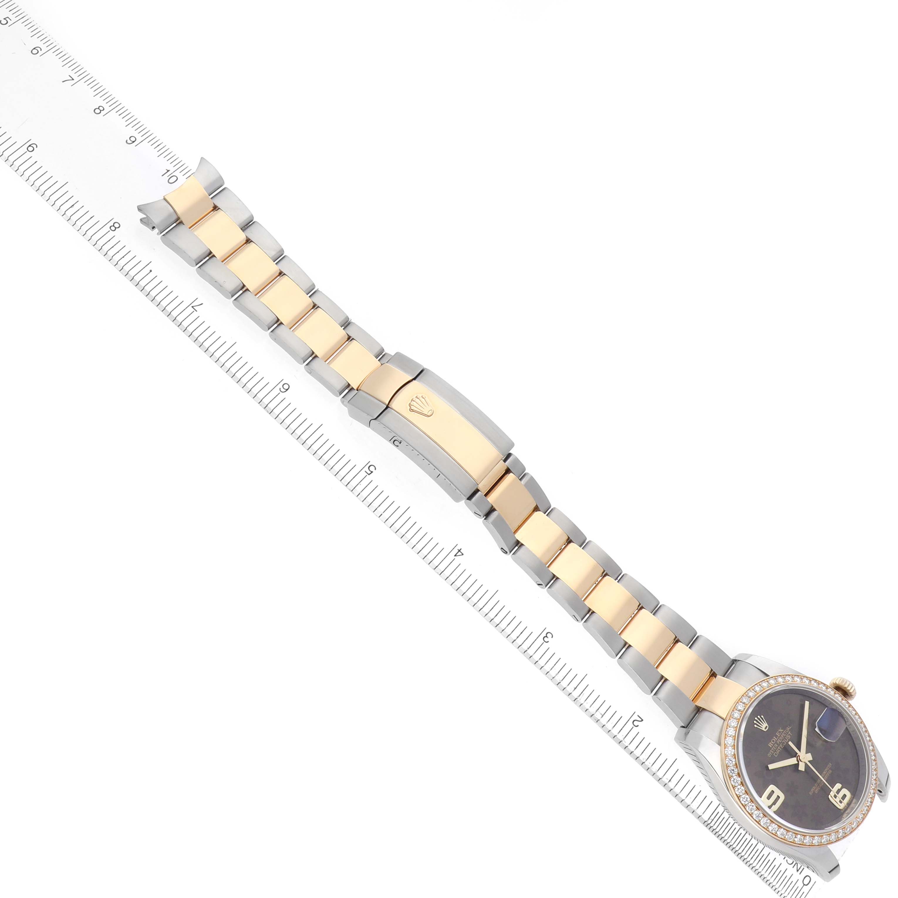 The Rolex Datejust 116243 Men's Steel and Gold (two tone) Brown Dial is shown from an overhead angle, featuring its two-tone bracelet and face alongside a ruler for scale.
