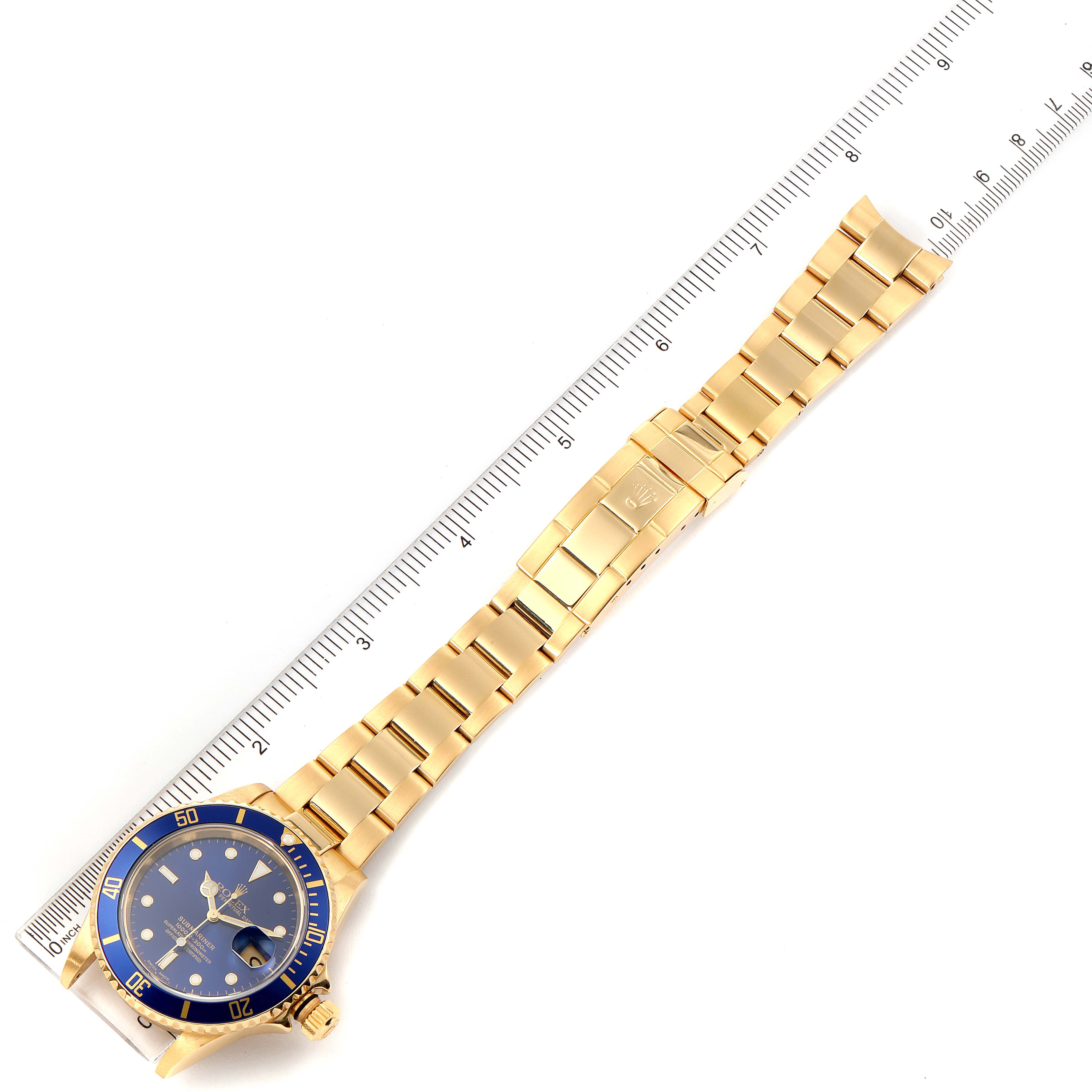 The image shows a top-down view of a Rolex Submariner 16618 Men
s Yellow Gold Blue Dial 16618 Men
s Yellow Gold Blue Dial watch, displaying its blue dial, bezel, and gold bracelet alongside a ruler.