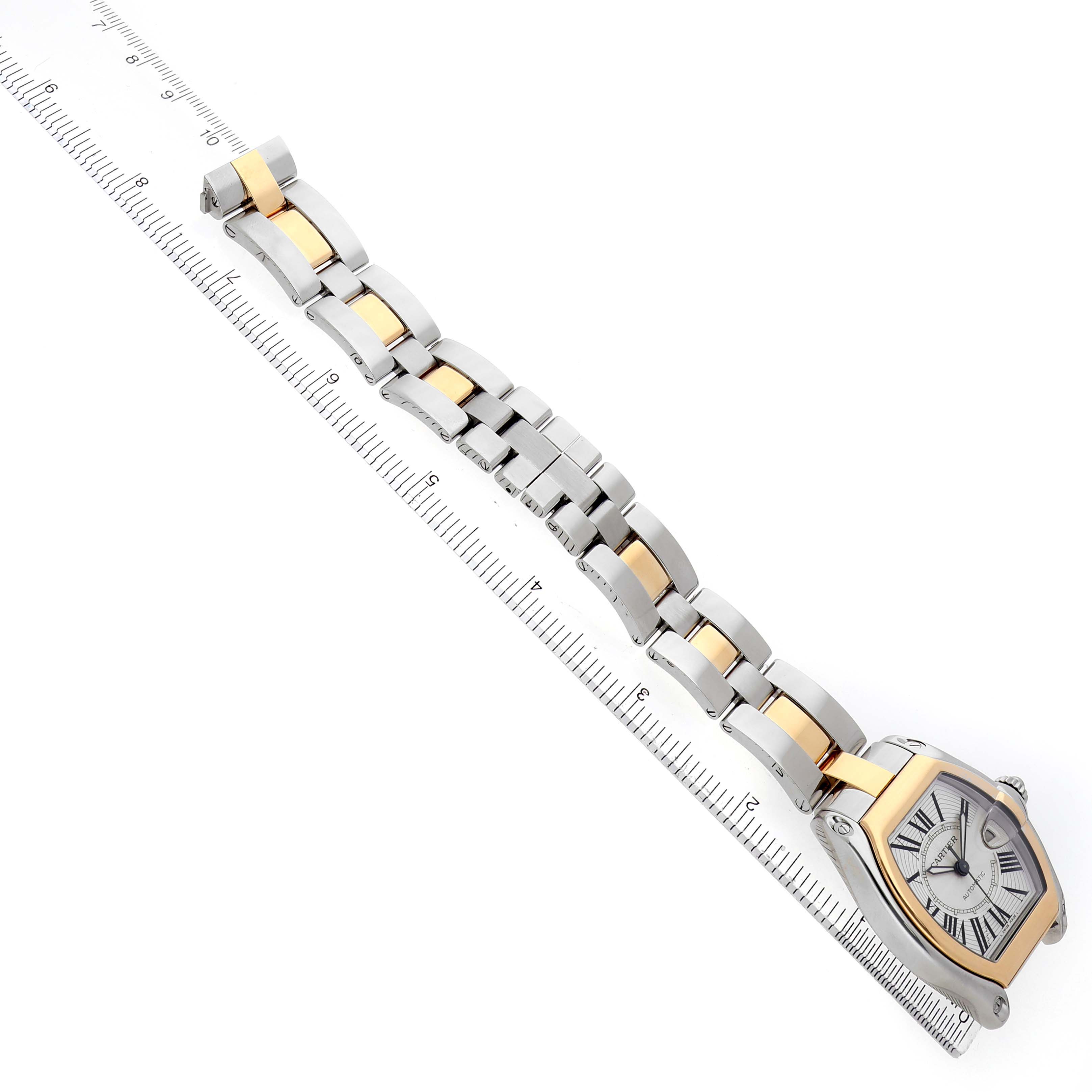 The Cartier Roadster W62031Y4 Men's Steel and Gold (two tone) Silver Dial watch is shown at a diagonal angle on a ruler, displaying its face and two-tone bracelet.