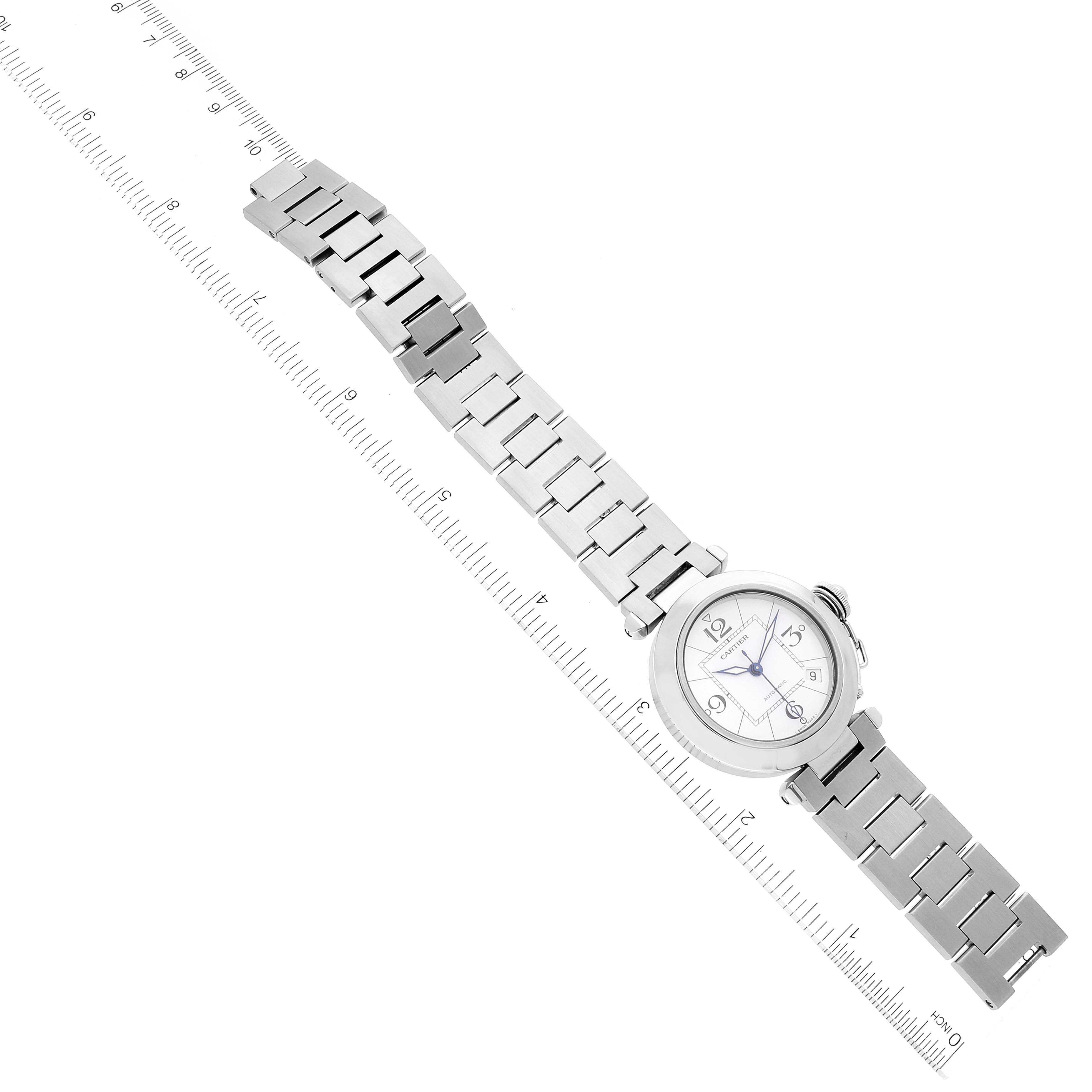 The Pasha model by Cartier is shown from a top-down angle, displaying the watch face, band, and crown next to a ruler for scale.