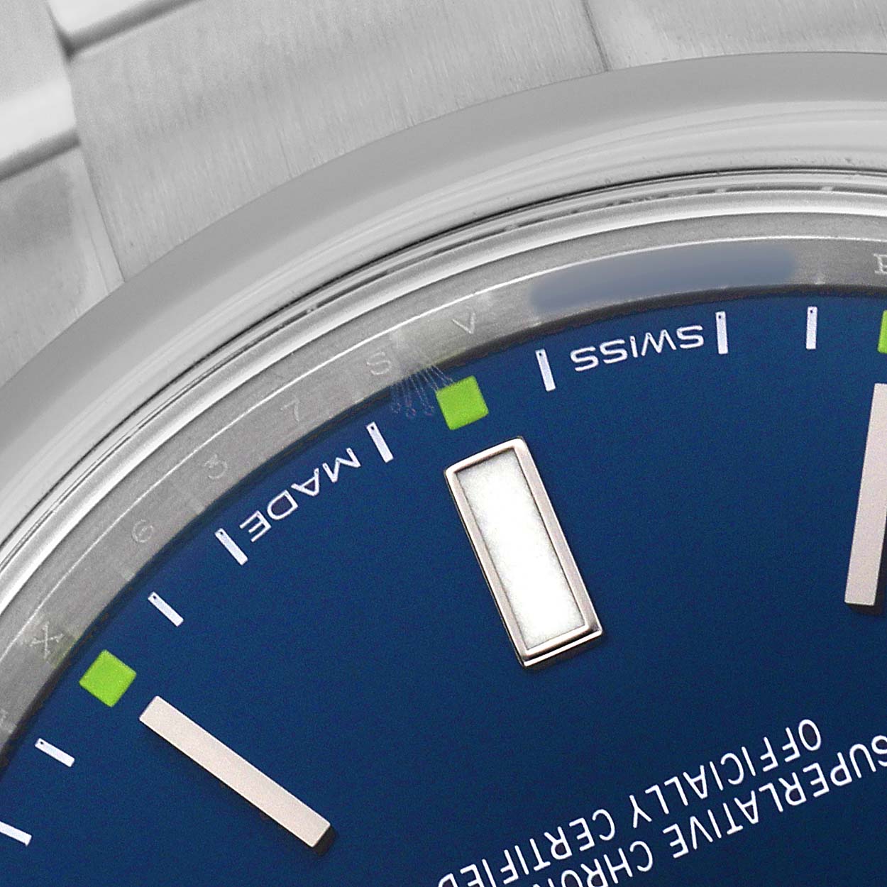 The image shows a close-up of the Rolex Oyster Perpetual 114300 Men's Stainless Steel Blue Dial watch, highlighting part of the blue dial and markers with a brushed bezel.