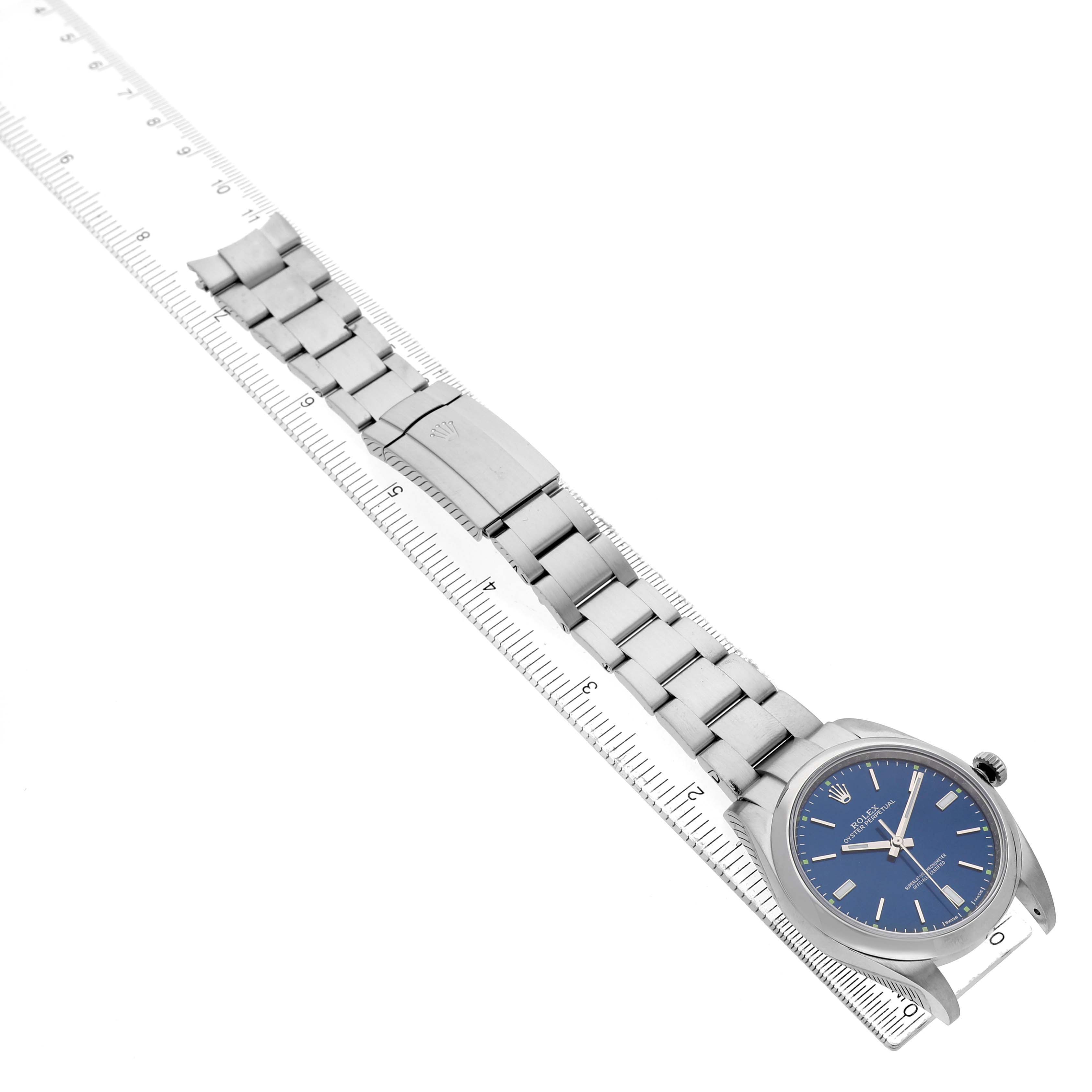 The Rolex Oyster Perpetual 114300 Men's Stainless Steel Blue Dial watch is shown at an angle displaying the face, bracelet, and clasp, positioned alongside a ruler.