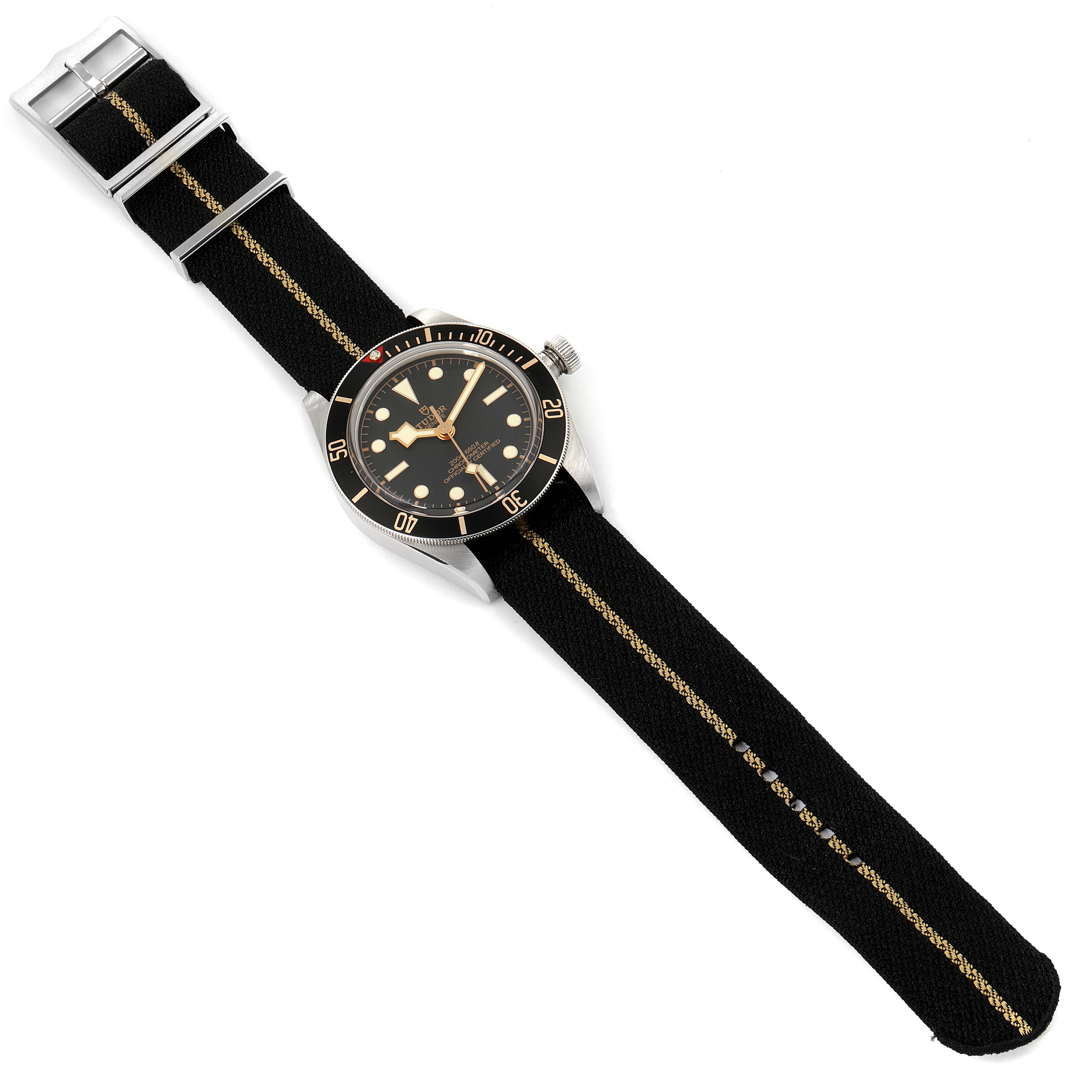 The image shows a Tudor Heritage Black Bay 79030N Men
s Stainless Steel Black Dial 79030N Men
s Stainless Steel Black Dial watch from a top-down angle, displaying its face and black NATO strap with a gold stripe.