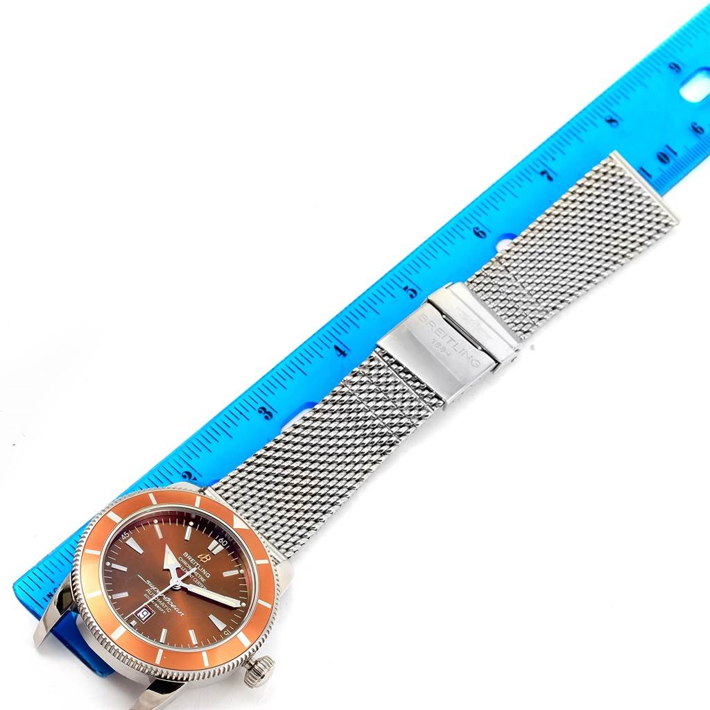 The image shows a Breitling Superocean A17320 Men's Stainless Steel Brown Dial A17320 Men's Stainless Steel Brown Dial watch with an orange bezel, mesh strap, and clasp, measured against a blue ruler.