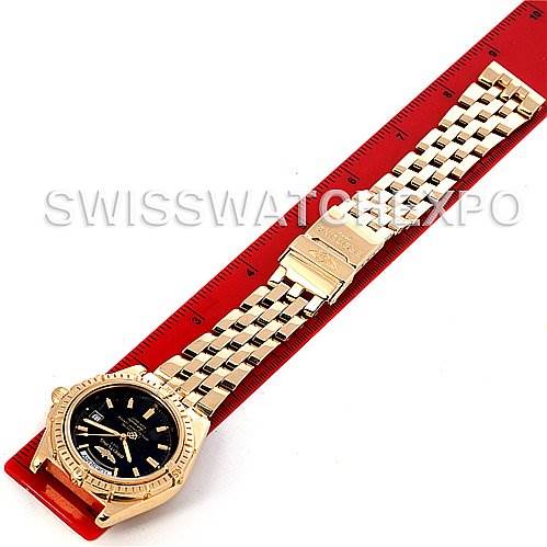 The image shows a Breitling Windrider Men's Yellow Gold Black Dial Men's Yellow Gold Black Dial watch placed horizontally on a red ruler, displaying the entire watch face and bracelet.